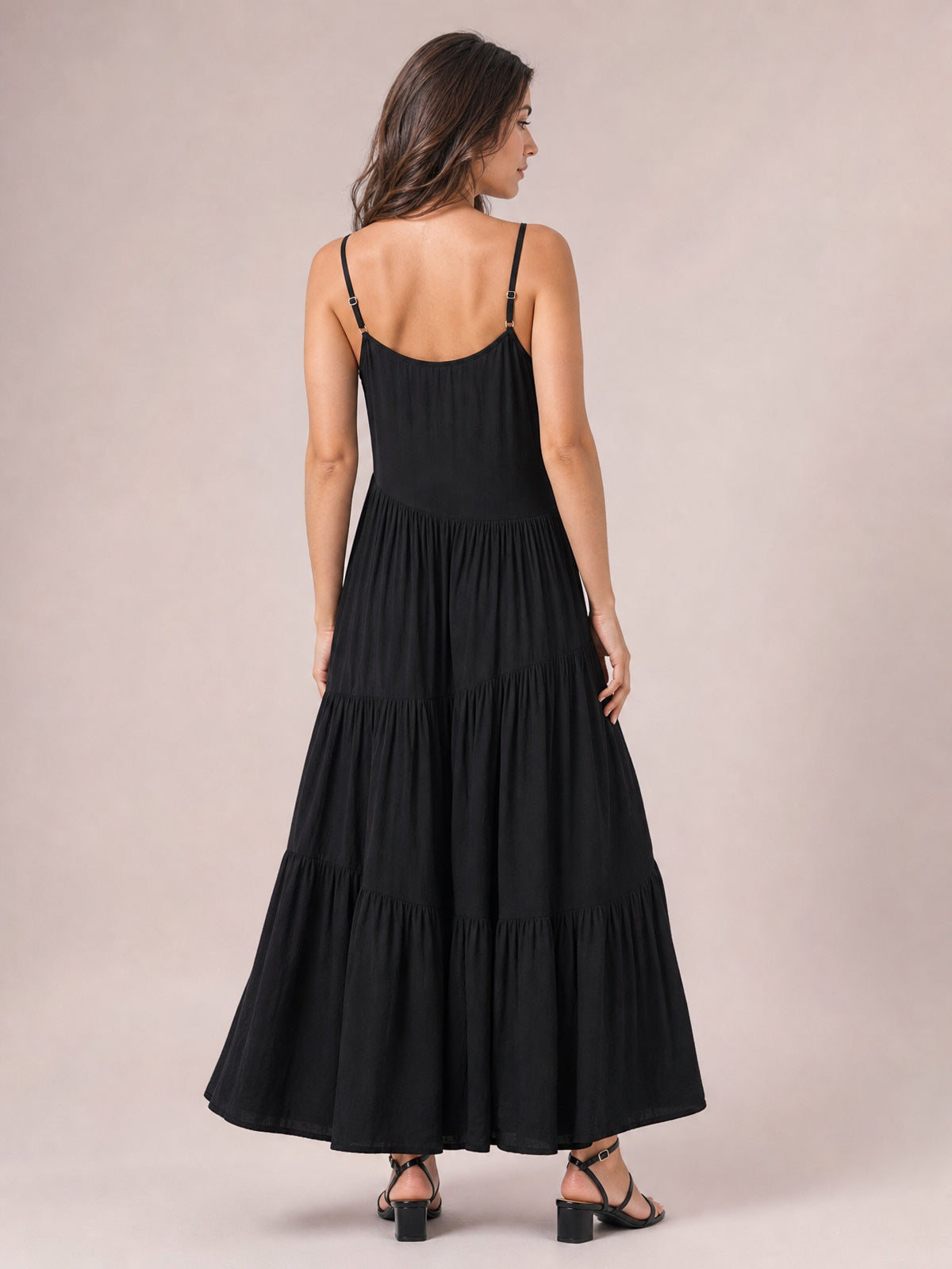Model shown from behind wearing a black ruched tiered spaghetti strap maxi dress with adjustable straps, a flowing full skirt, and strappy heels.