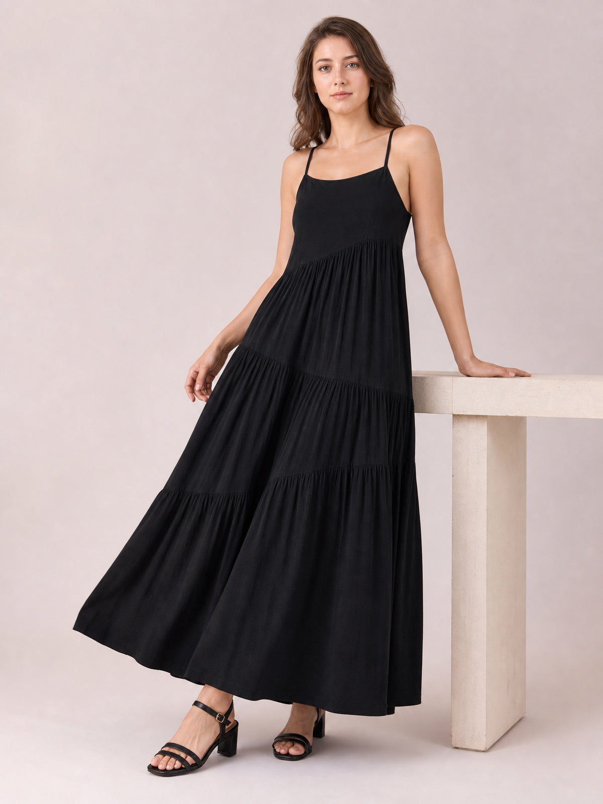 Model poses in a black ruched tiered spaghetti strap maxi dress with a scoop neckline and flowing, full silhouette.