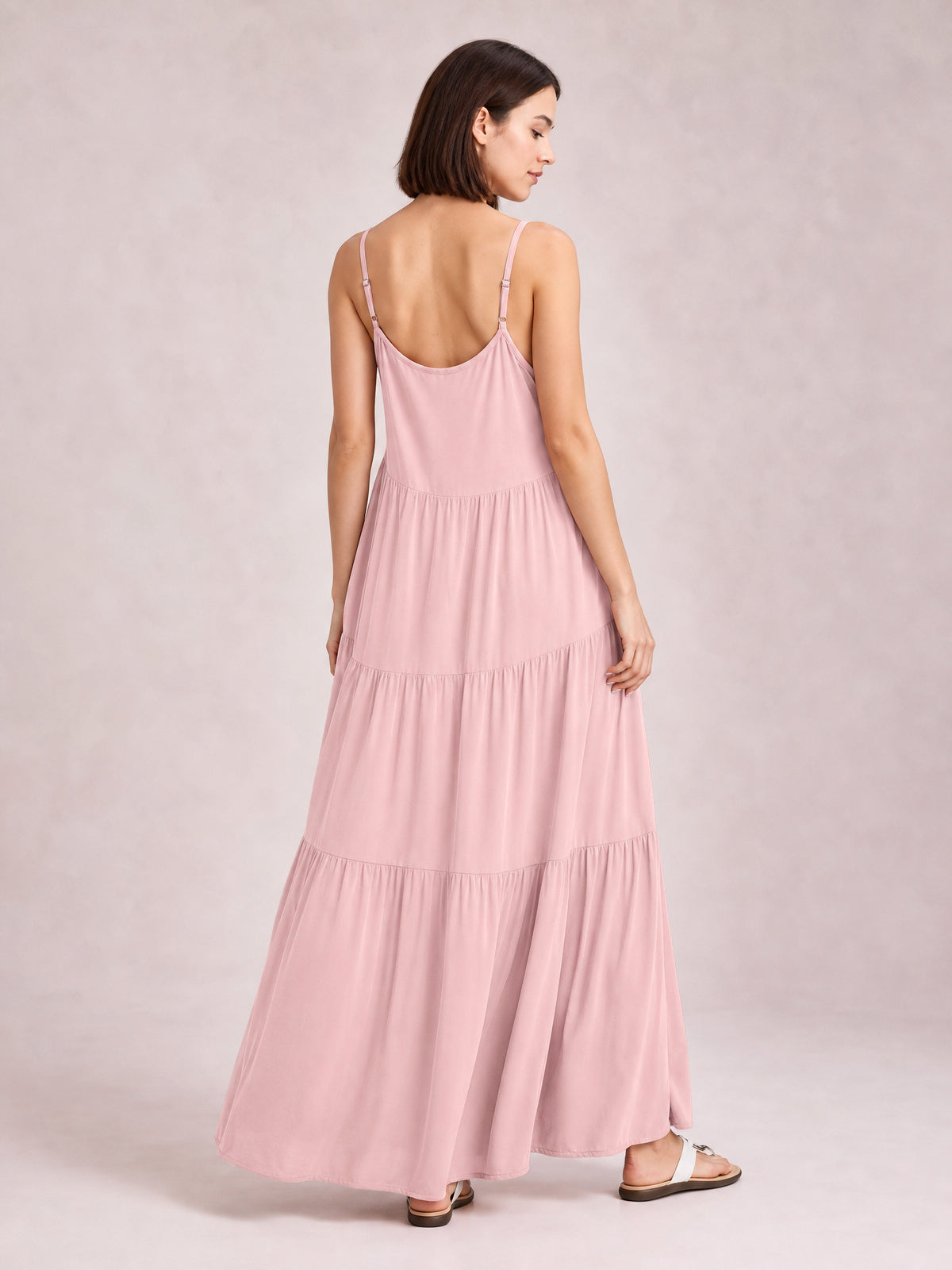 Model turned away wearing a blush pink ruched tiered spaghetti strap maxi dress with a flowing floor-length silhouette and softly gathered skirt.