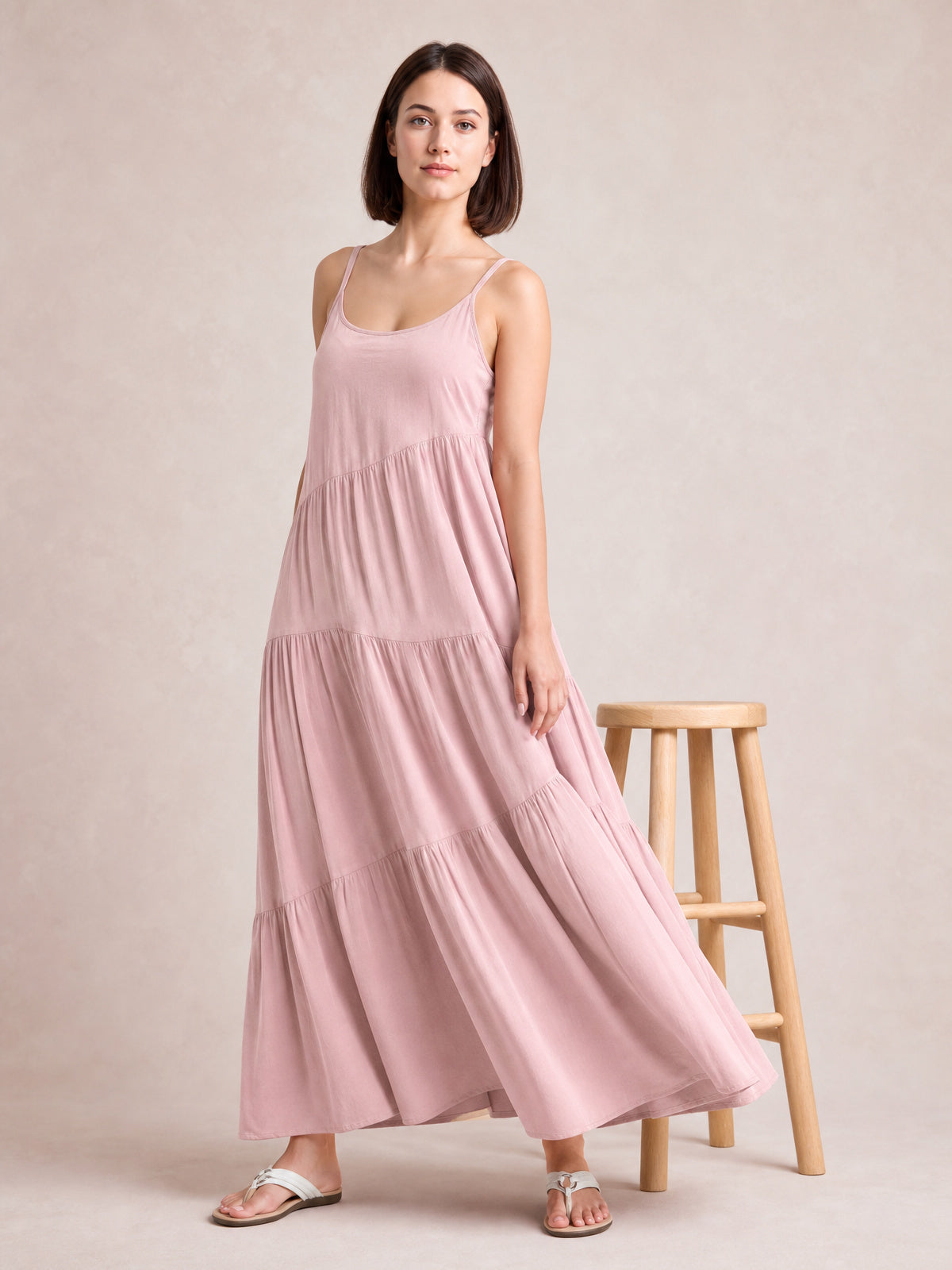 Model poses in a flowing pink ruched tiered spaghetti strap maxi dress with a scoop neckline, gathered seams, and full swaying skirt.