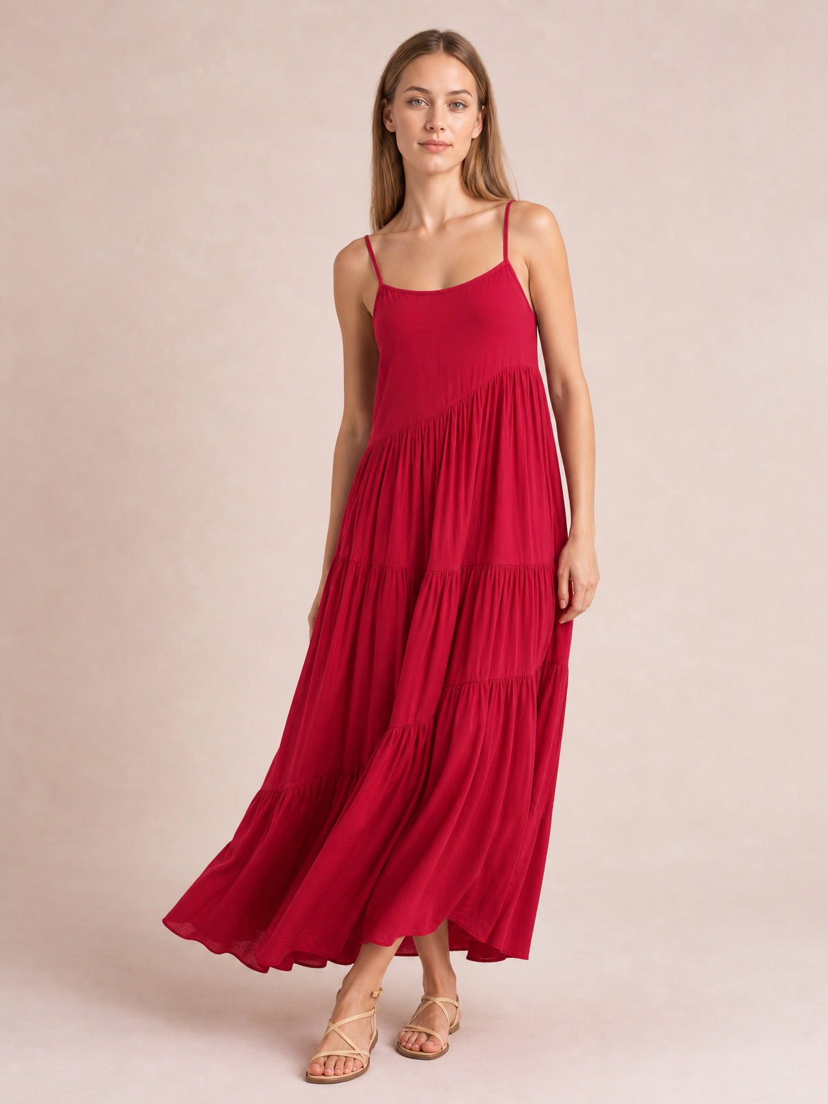 Model poses in a red ruched tiered spaghetti strap maxi dress with a scoop neckline, gathered skirt, flowing full silhouette, and strappy sandals.