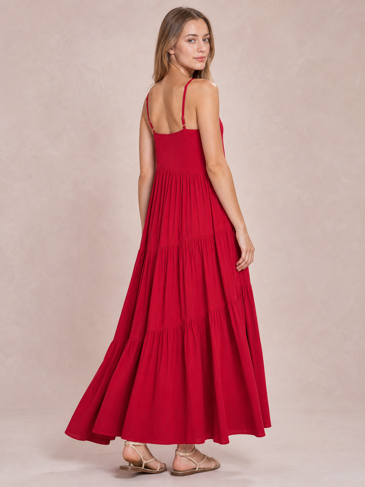 Model poses in a red ruched tiered spaghetti strap maxi dress with a fitted bodice, adjustable straps, gathered panels, and a flowing floor-length silhouette.