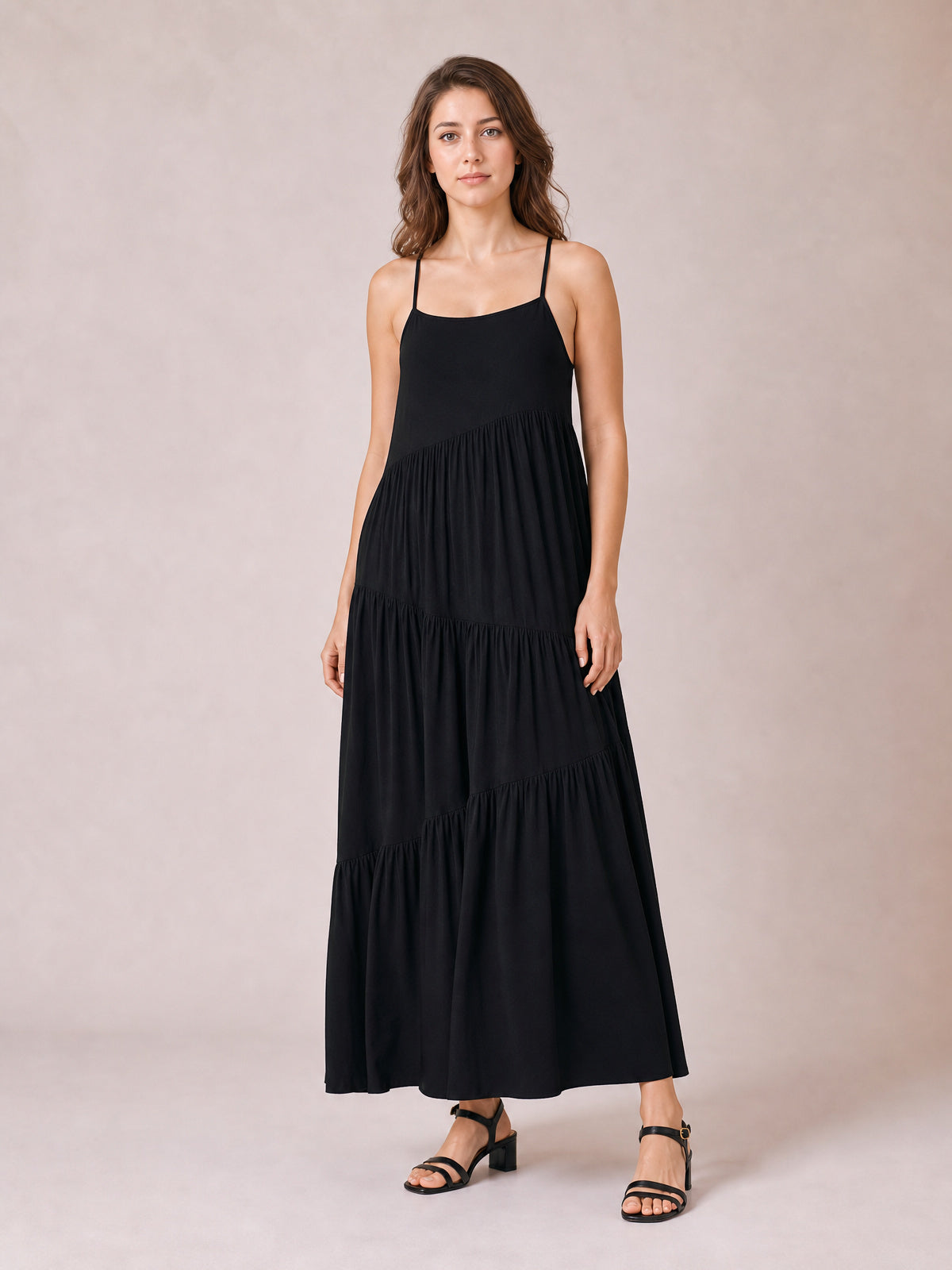 Model stands in a black ruched tiered spaghetti strap maxi dress with a scoop neckline, flowing full silhouette, and floor-skimming hem.
