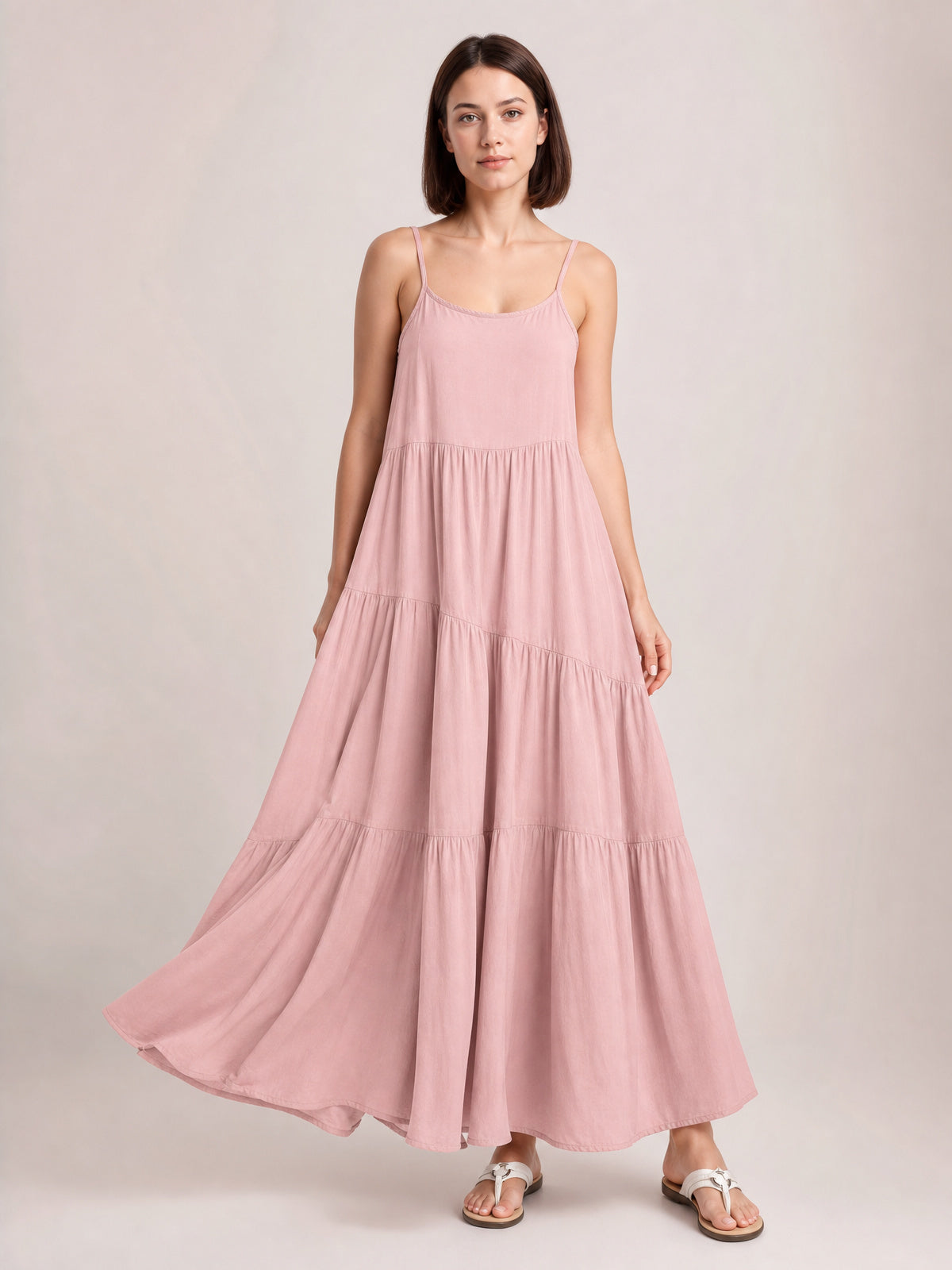 Model poses in a flowing blush pink ruched tiered spaghetti strap maxi dress with a scoop neckline, gathered seams, and full floor-skimming skirt.