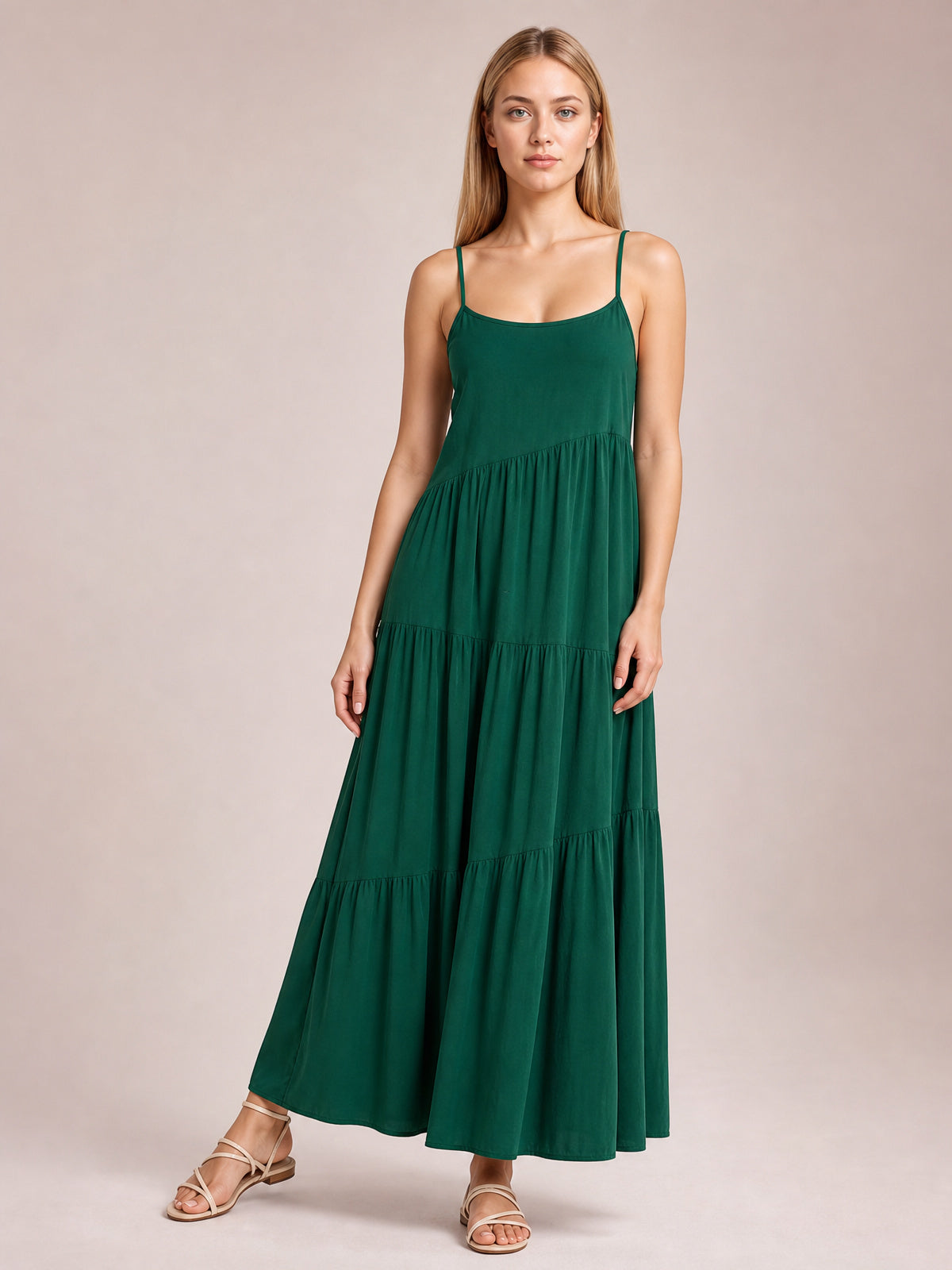 A model poses in a deep green ruched tiered maxi dress with slim spaghetti straps, a scoop neckline, and a flowing full-length silhouette with gathered skirt panels.