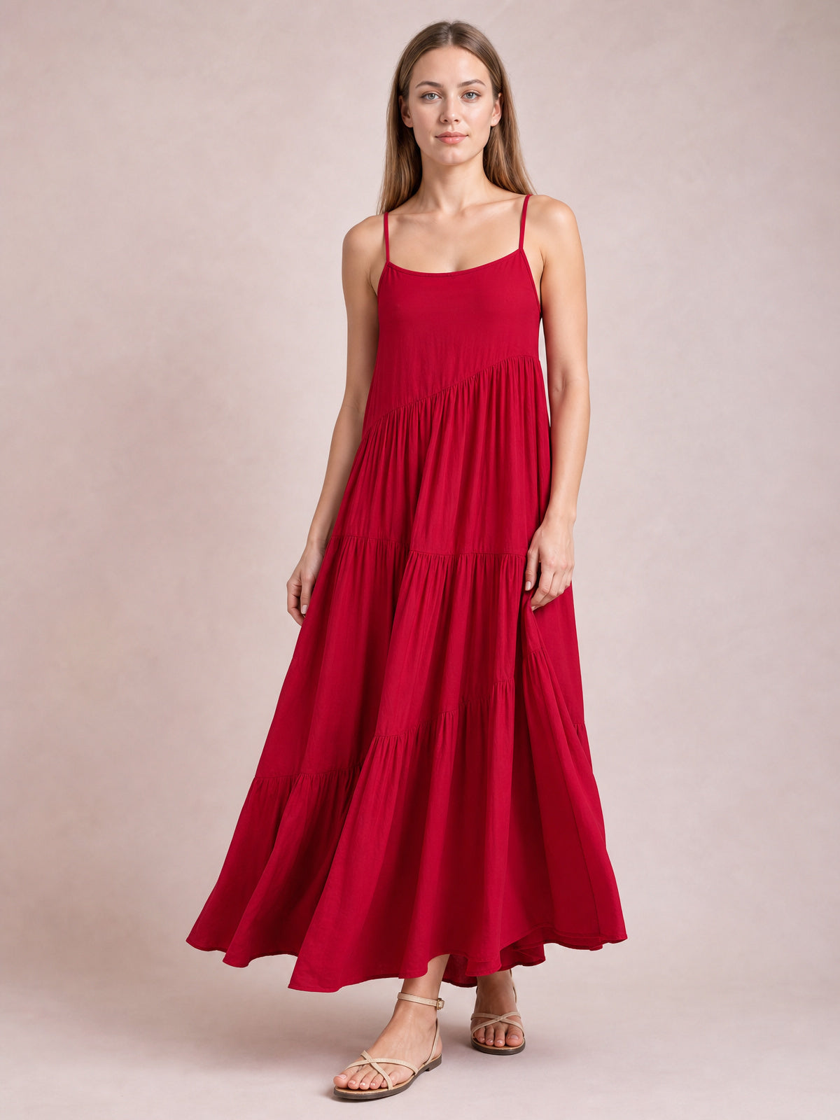 Model poses in a red ruched tiered spaghetti strap maxi dress with a scoop neckline, flowing full silhouette, and floor-skimming hem.