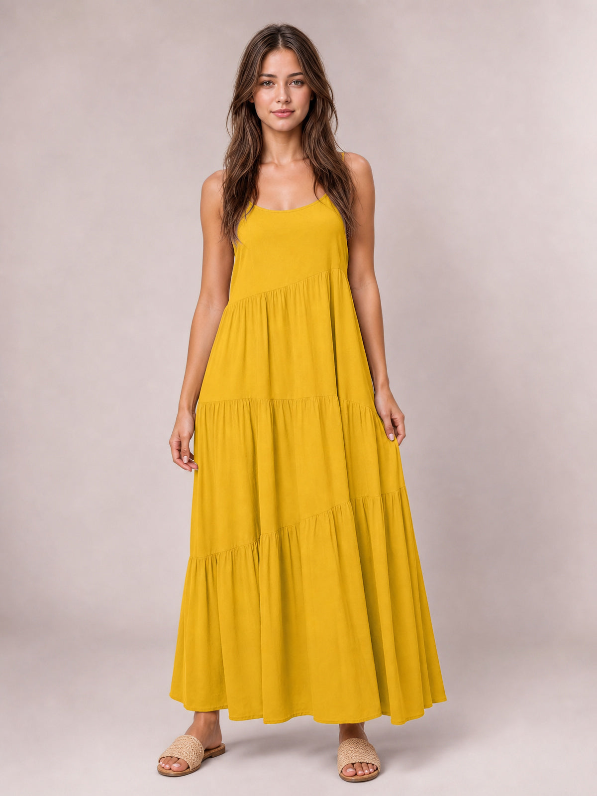 A model stands in a mustard yellow spaghetti strap maxi dress with a scoop neckline, gathered tiered skirt, flowing silhouette, and hands holding the sides.