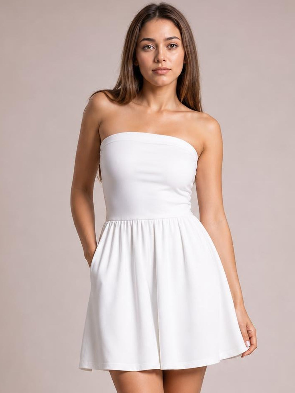 A model wears a white ruched tube mini dress with a strapless fitted bodice, gathered waist, and flared short skirt, posing with one hand in a pocket.