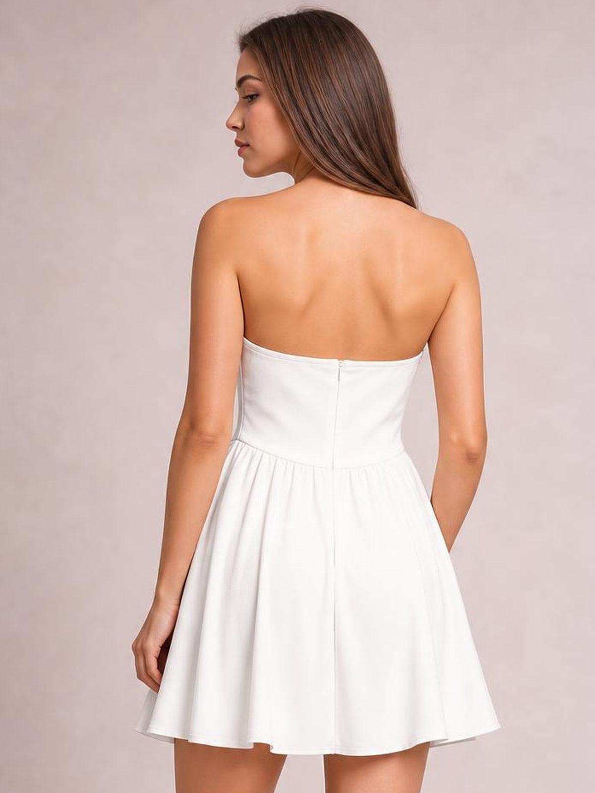 Back view of a model wearing a white strapless mini dress with a fitted bodice, center-back zipper, and flared skirt, standing with one hand in a pocket.