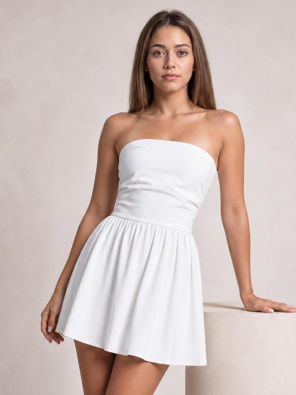 A model wears a white ruched tube mini dress with a strapless neckline, fitted bodice, gathered waist, and flared short skirt while leaning against a ledge.
