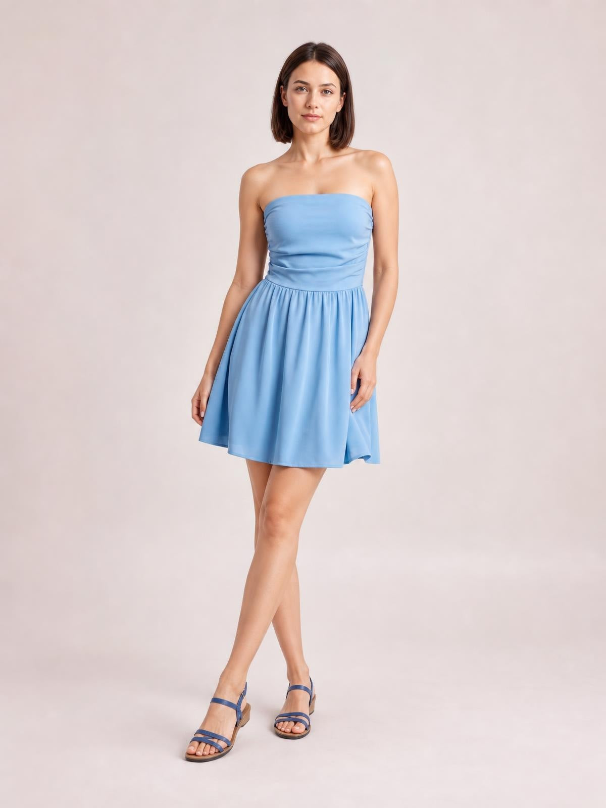 A model poses in a light blue ruched tube mini dress with a strapless bodice, gathered waist, and flared skirt, crossing her legs.
