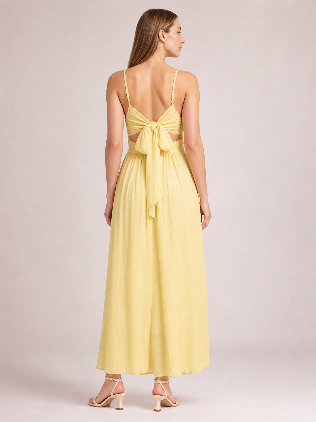 Model shown from behind in a pale yellow cami dress with slender straps, a tied ruched back, gathered waist, and flowing floor-length skirt.