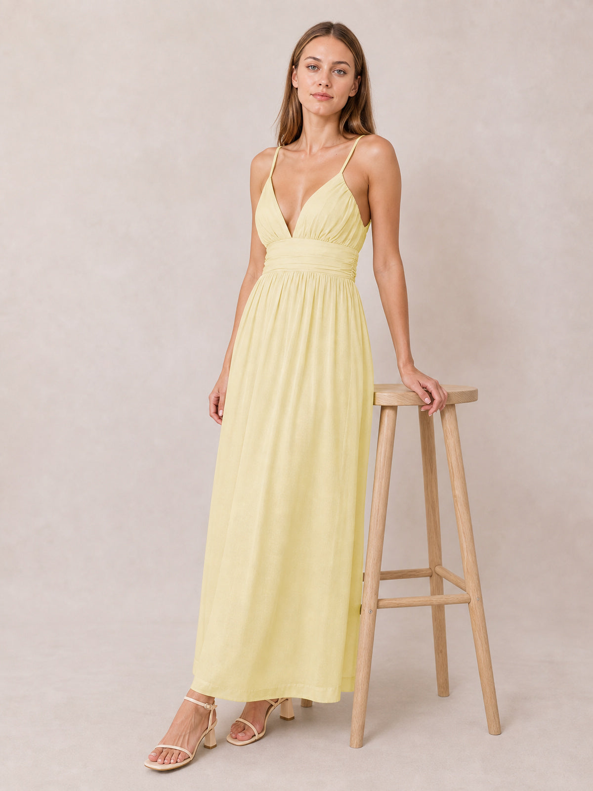 A model poses beside a stool in a pale yellow ruched V-neck cami dress with slender straps, gathered bodice, defined waist, and flowing maxi silhouette.