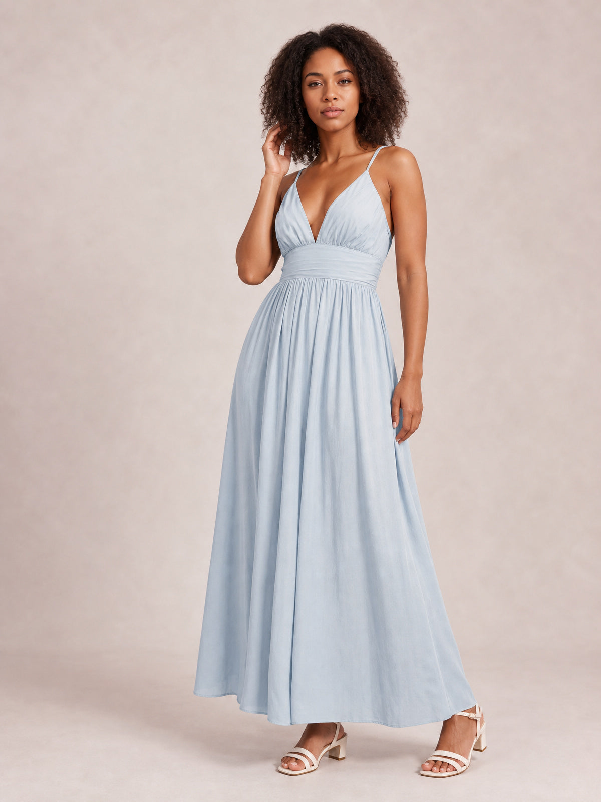 Model in a light blue ruched V-neck cami dress with slender straps, a defined waist, and a flowing floor-length skirt, posing with one hand near her hair.