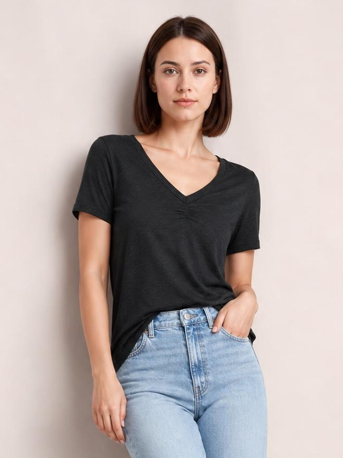 Model wearing a black ruched V-neck short sleeve T-shirt with a relaxed silhouette, standing with one hand in her jeans pocket.