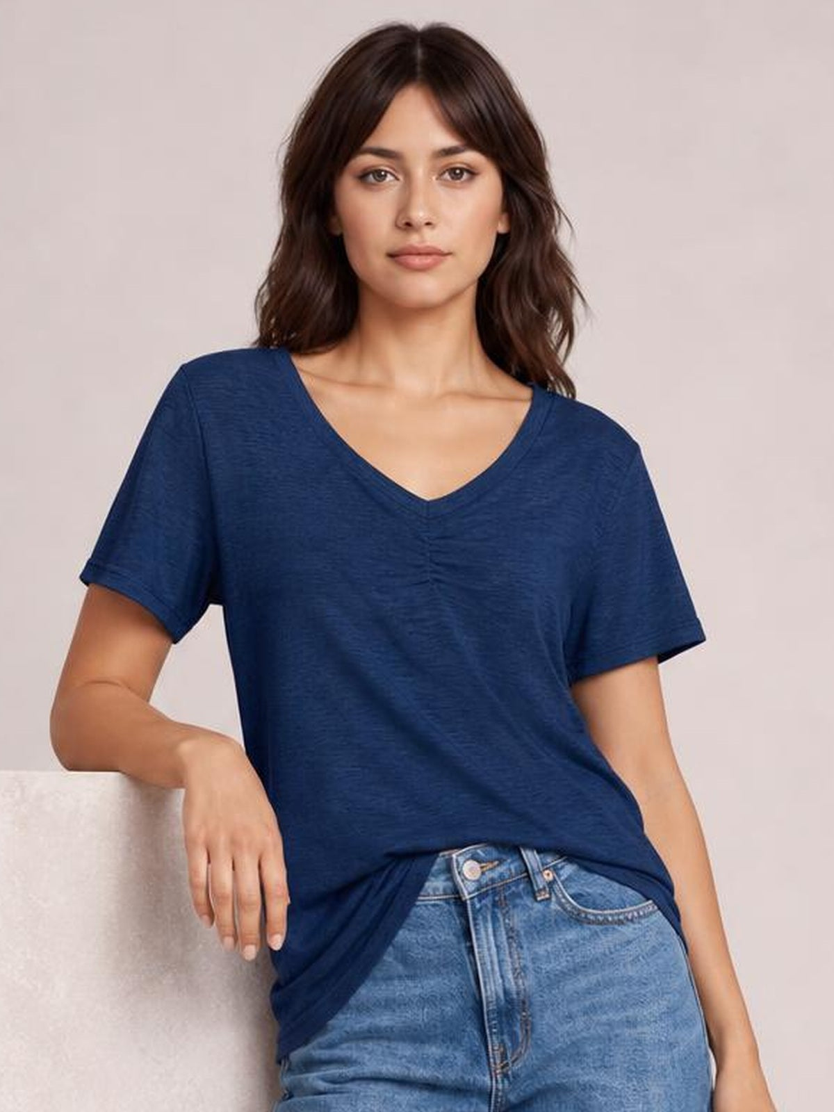Model leans against a surface wearing a navy blue ruched V-neck short sleeve T-shirt with a relaxed silhouette and gathered center detail.