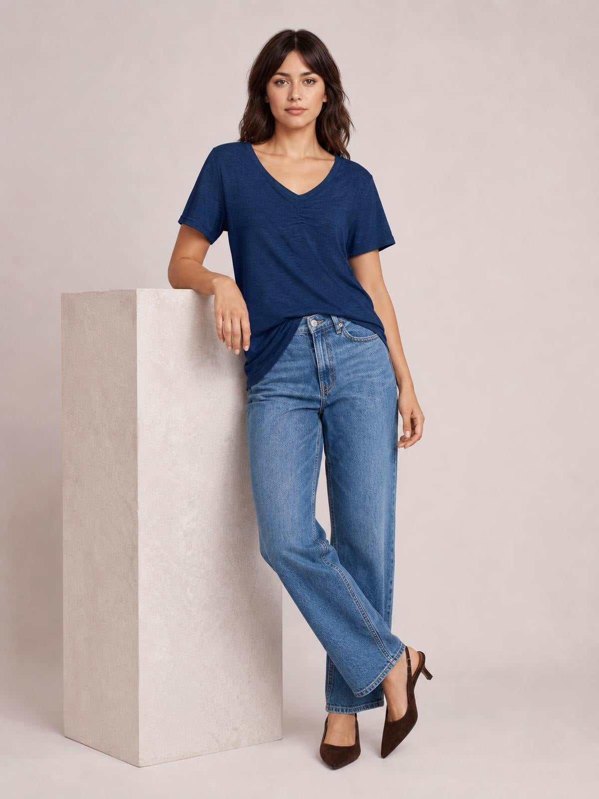 Model wearing a navy ruched V-neck short sleeve T-shirt with a relaxed silhouette, paired with jeans while leaning casually against a pedestal.