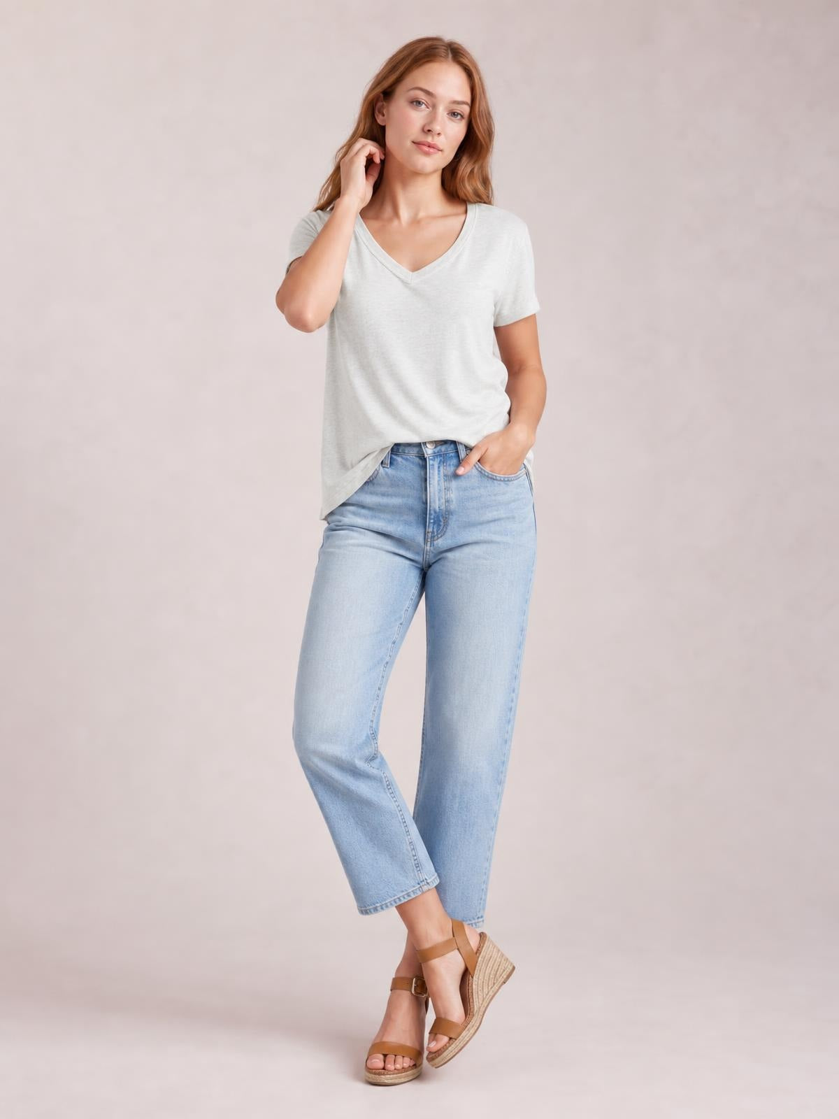 A model wears a light gray V-neck short-sleeve T-shirt with a relaxed silhouette and subtle neckline ruching, standing with one hand in her jeans pocket.