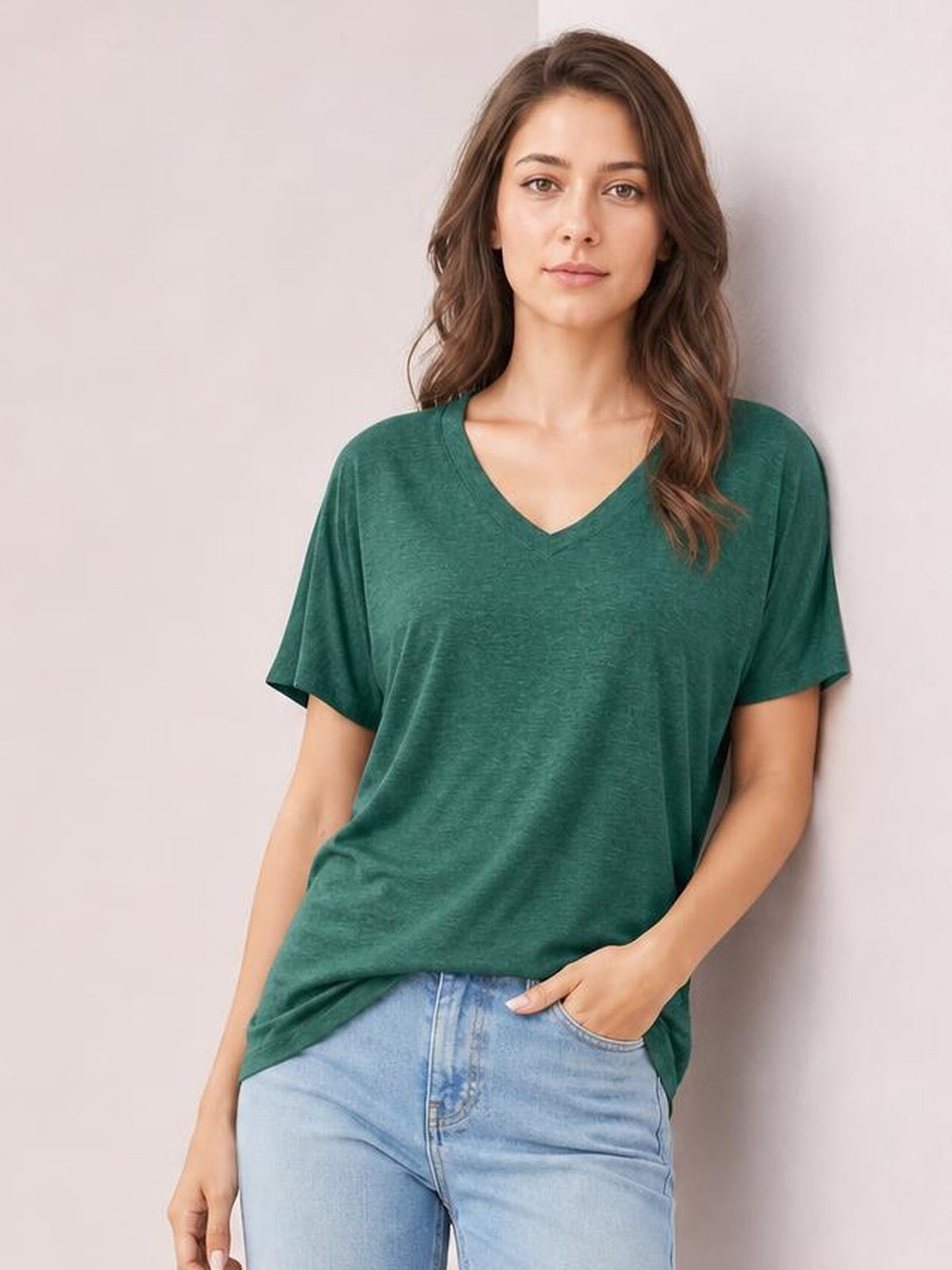 A model wears a soft green ruched V-neck short sleeve T-shirt with a relaxed, drapey silhouette, standing with one hand in her jeans pocket.