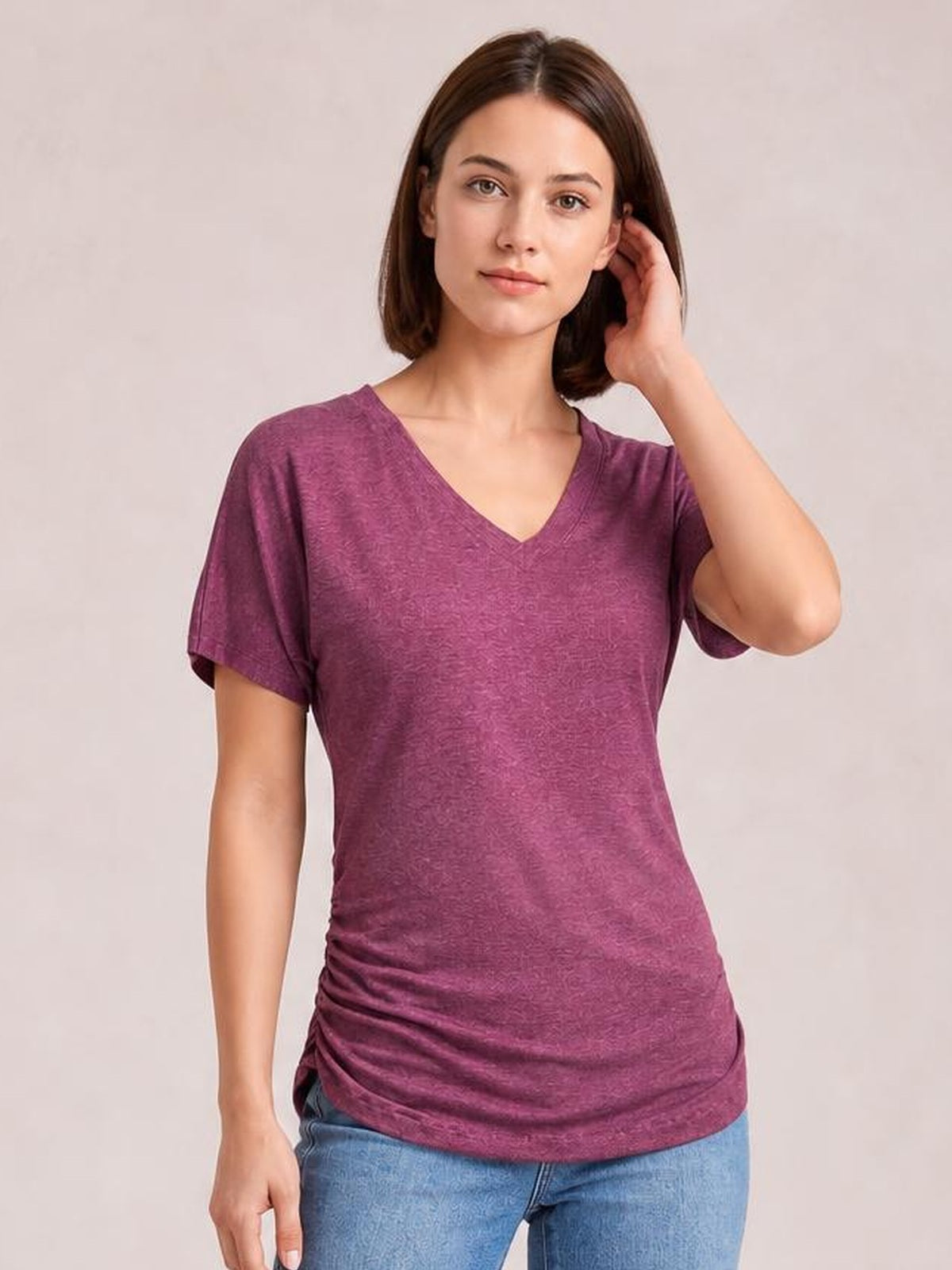 Model wearing a burgundy ruched V-neck short sleeve T-shirt with a stretchy, fitted silhouette, standing with one hand touching her hair.