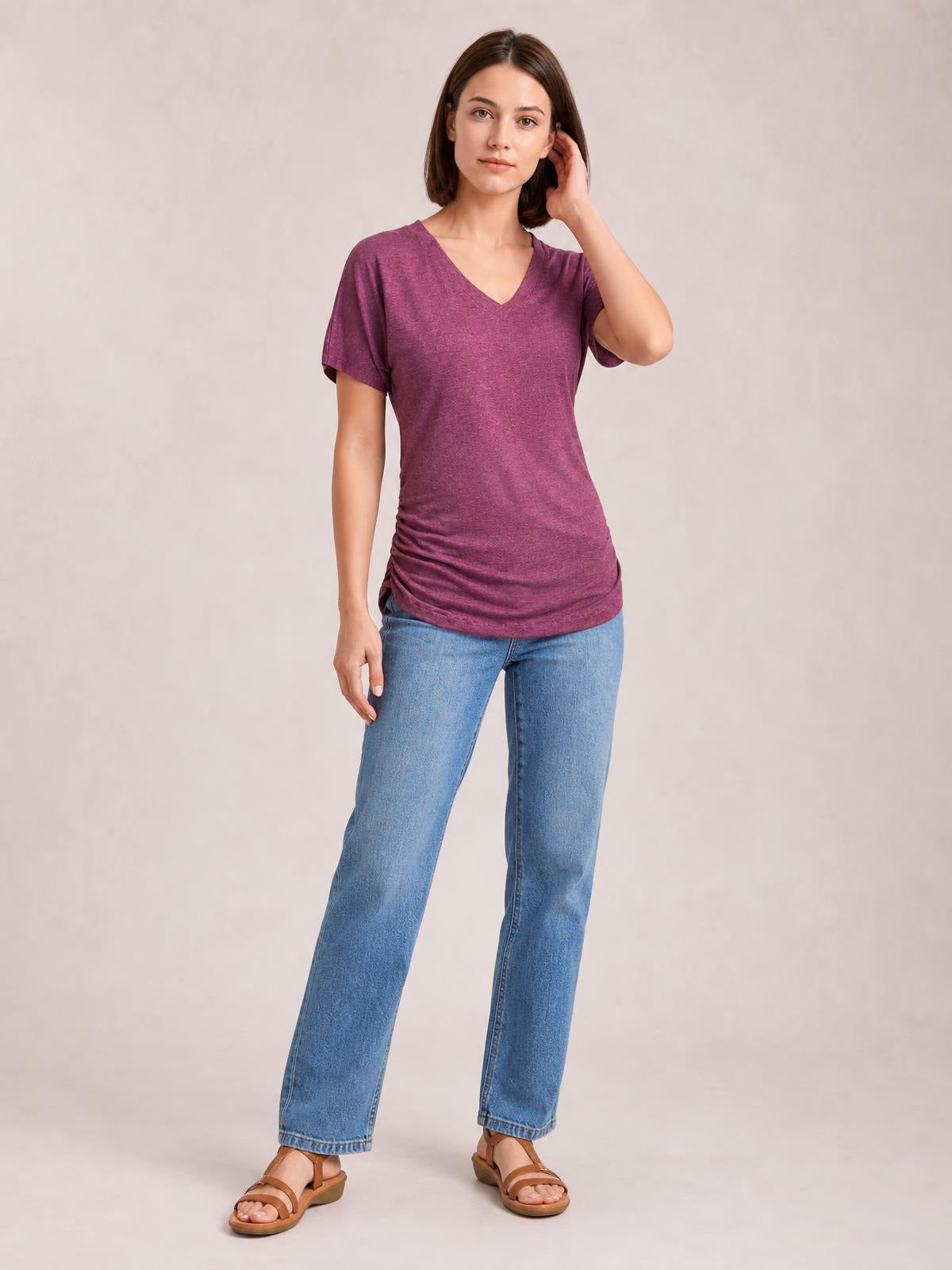 Model in a plum ruched V-neck short sleeve T-shirt with a fitted, stretchy silhouette and gathered side seams, standing with one hand touching her hair.