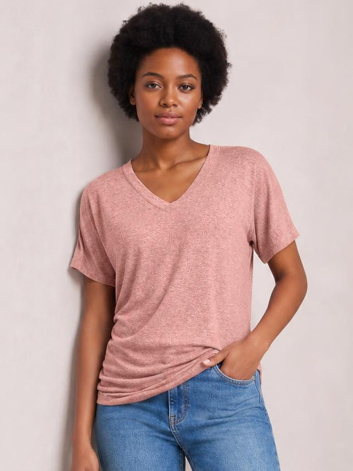 A model poses in a soft pink ruched V-neck short-sleeve T-shirt with a relaxed silhouette and gathered hem, one hand in her jeans pocket.