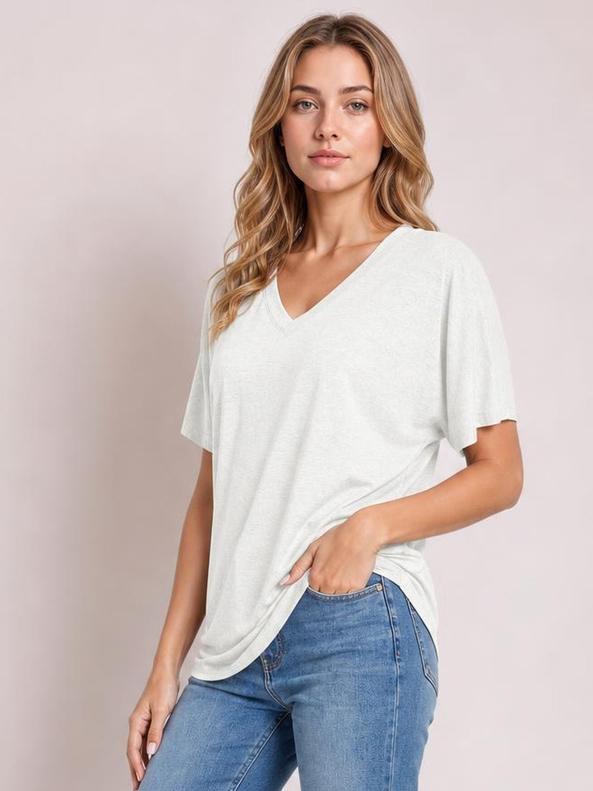 A model wears a white V-neck short-sleeve T-shirt with a relaxed silhouette, gently draped hem, and soft stretchy construction while posing with one hand at her jeans pocket.