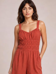 Model poses in a rust ruched jumpsuit with wide straps, a gathered bodice, wide-leg silhouette, and a visible side pocket.