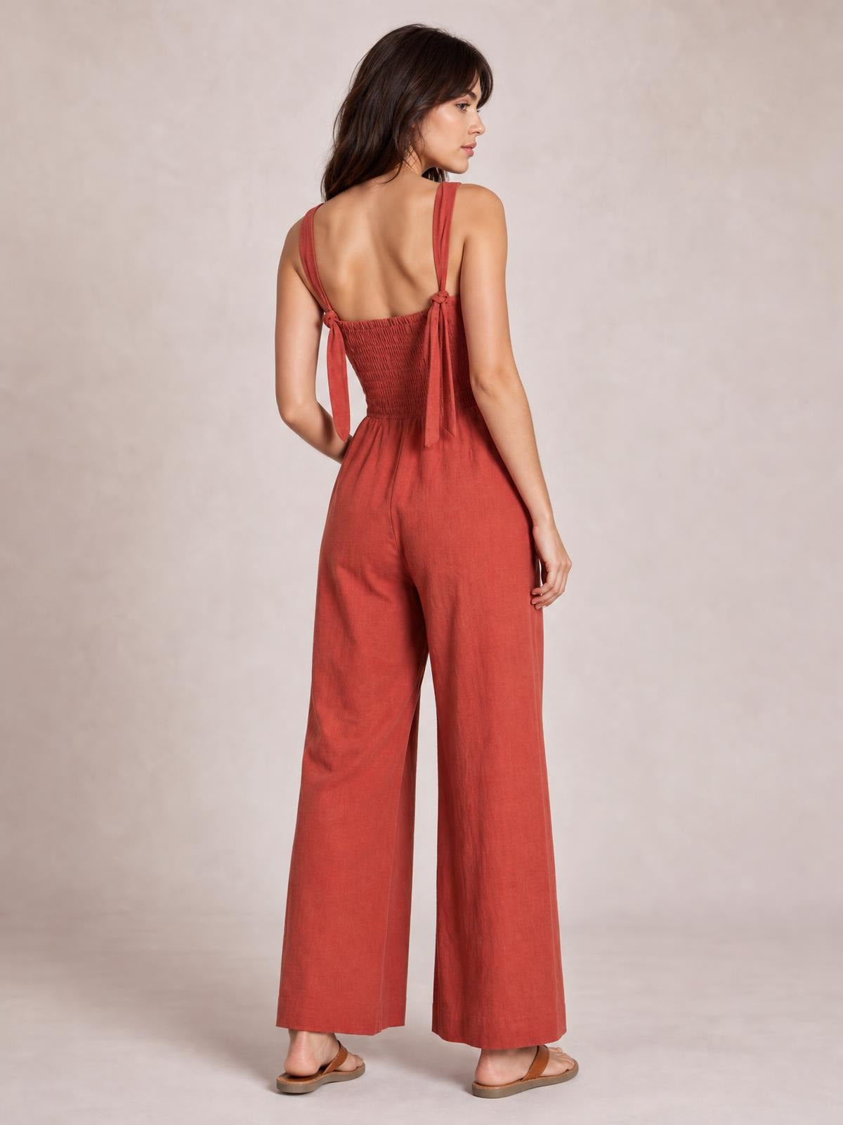 A model poses in a rust-red ruched wide strap jumpsuit, shown from the back with tied shoulder straps, a fitted smocked waist, and flowing wide legs.