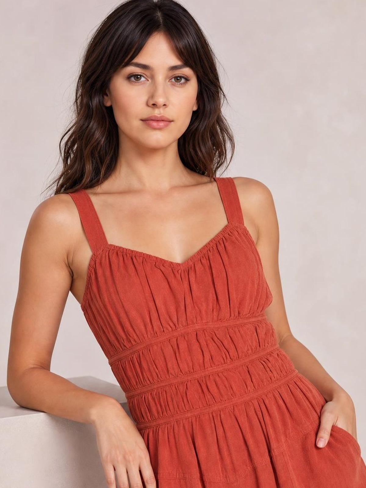 A model leans in a rust-red ruched jumpsuit with wide straps, a gathered bodice, and a defined waist for a relaxed silhouette.