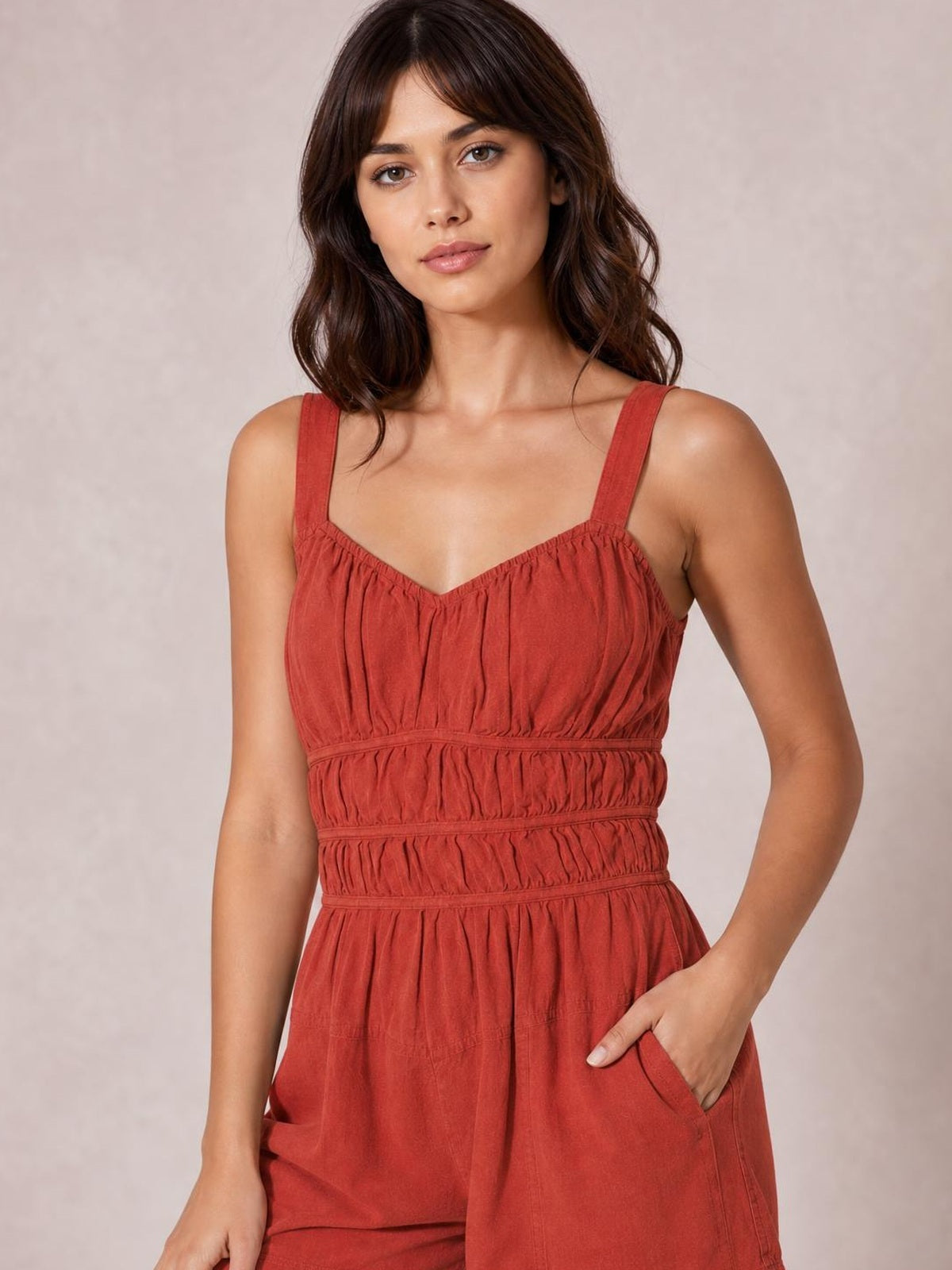 Model poses in a rust ruched jumpsuit with wide straps, a gathered bodice, defined waist, wide-leg silhouette, and visible side pockets.
