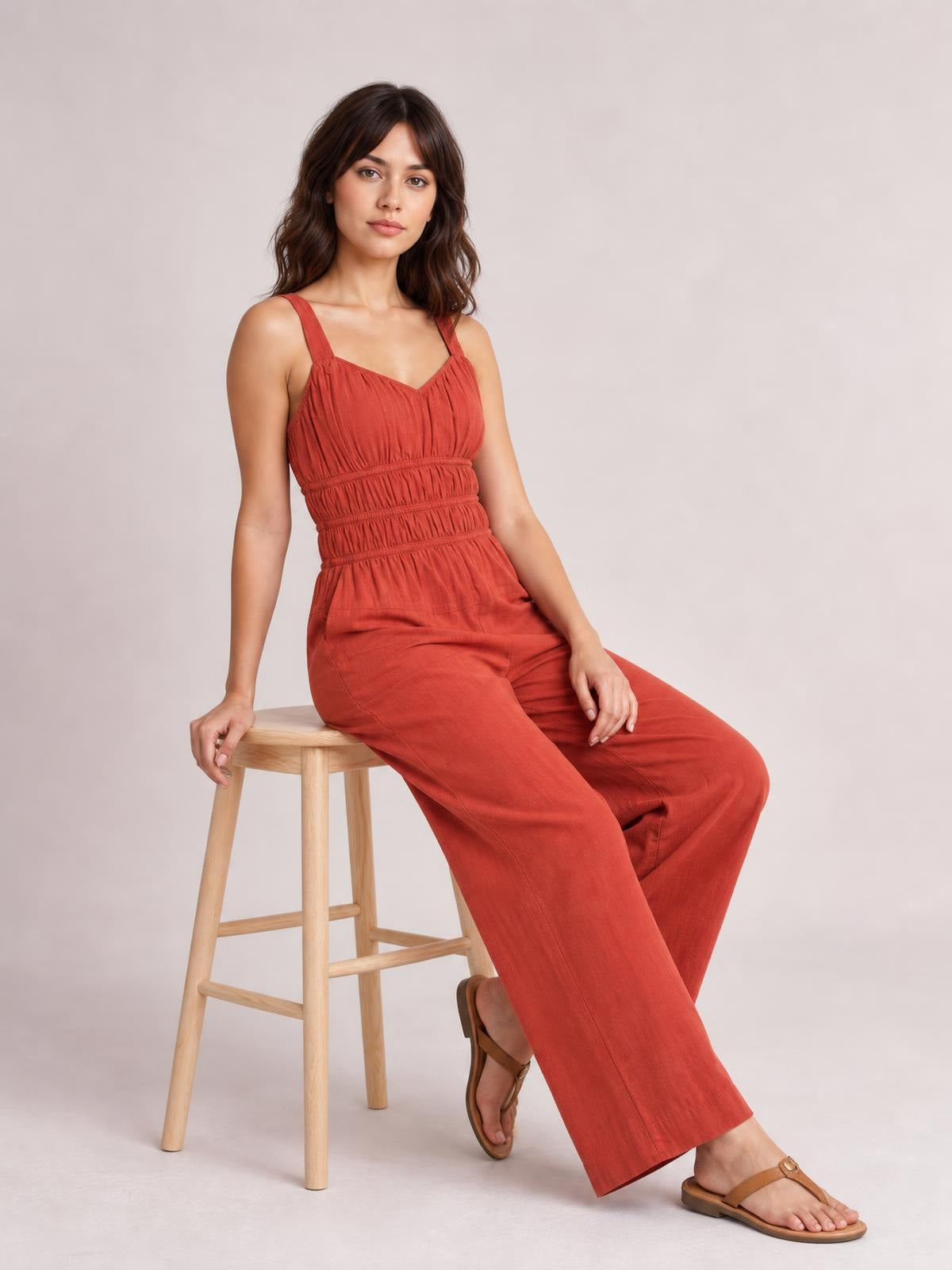 Model seated on a stool wears a rust-red ruched wide-strap jumpsuit with a tied neckline, smocked waist, pockets, and wide-leg silhouette.