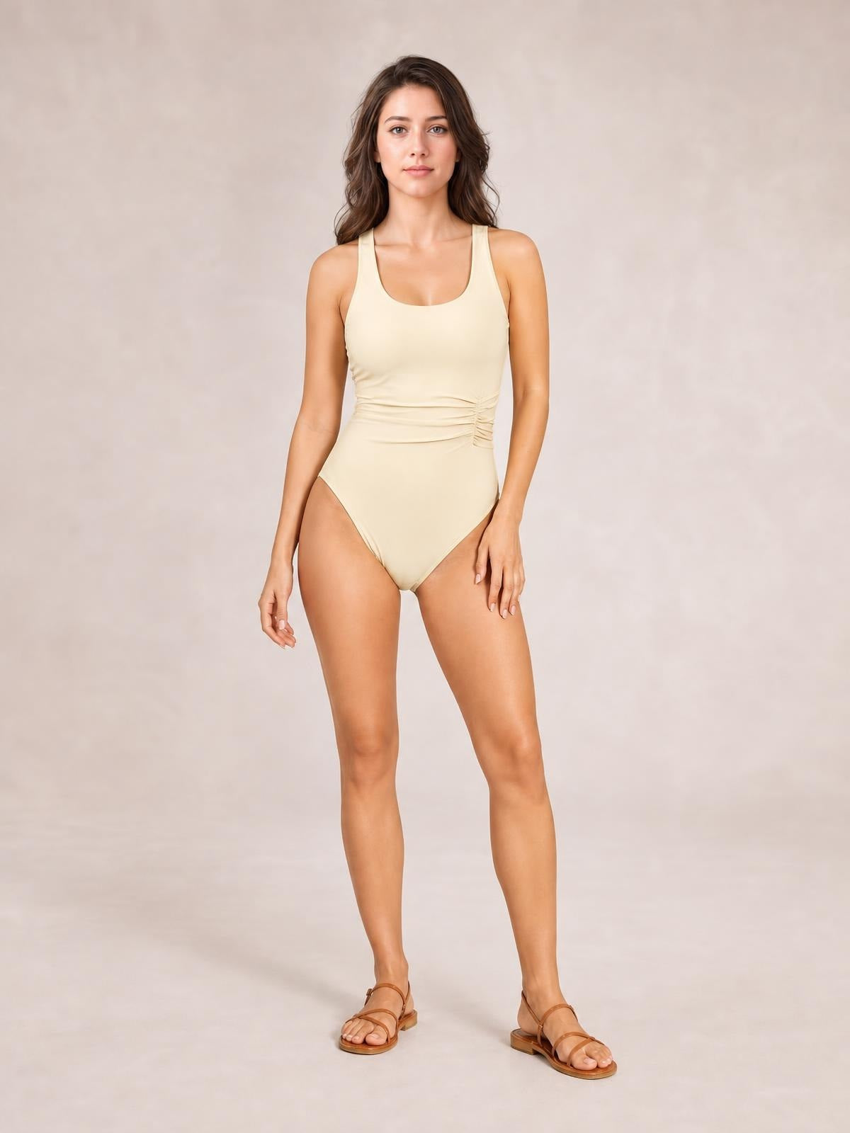 A model stands in a cream ruched one-piece swimsuit with wide straps, a scoop neckline, high-cut legs, and a fitted, sculpting silhouette.