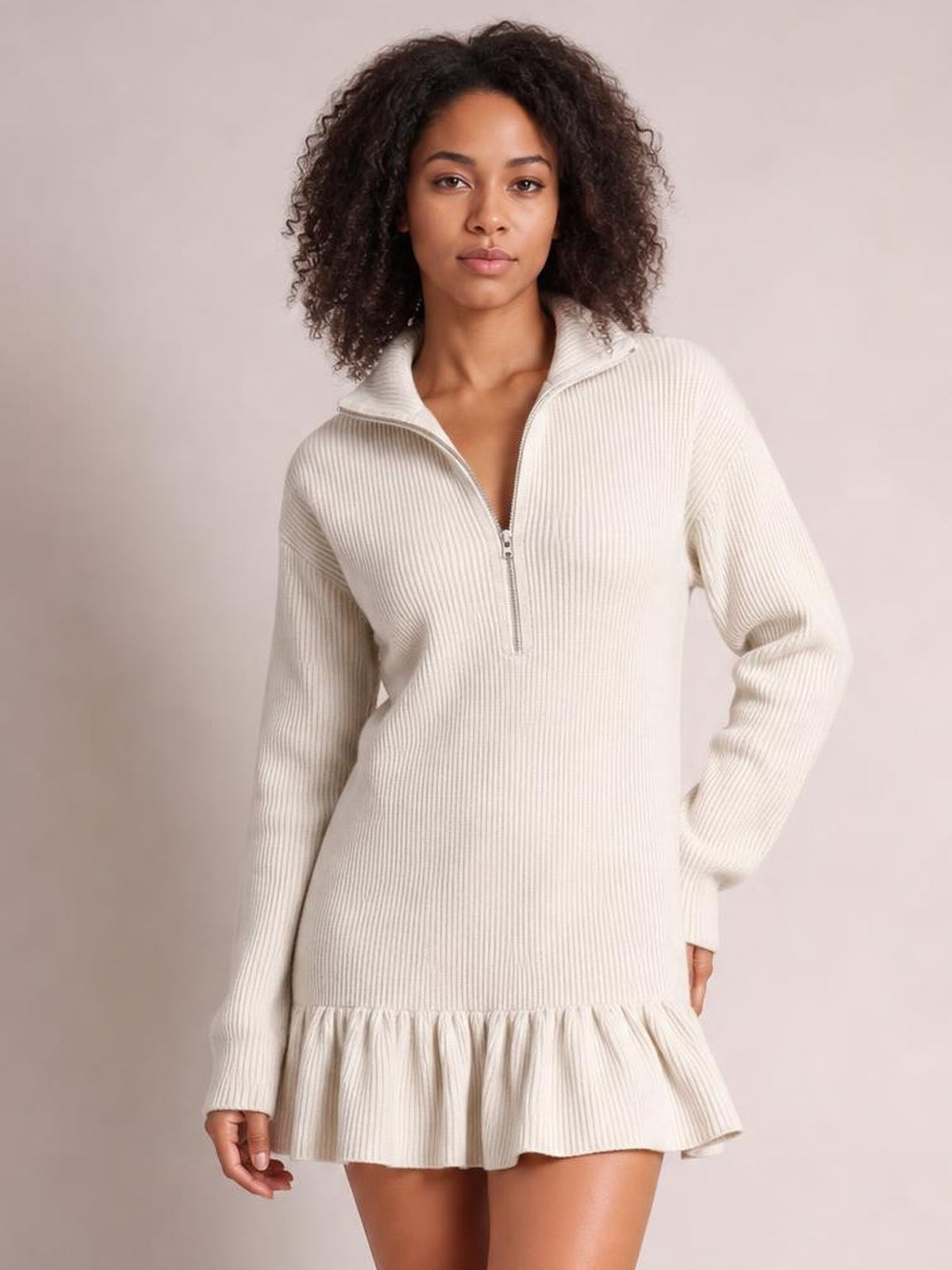 Model stands in a cream ribbed dress with long sleeves, a half-zip high collar, relaxed silhouette, and gathered ruffle hem.