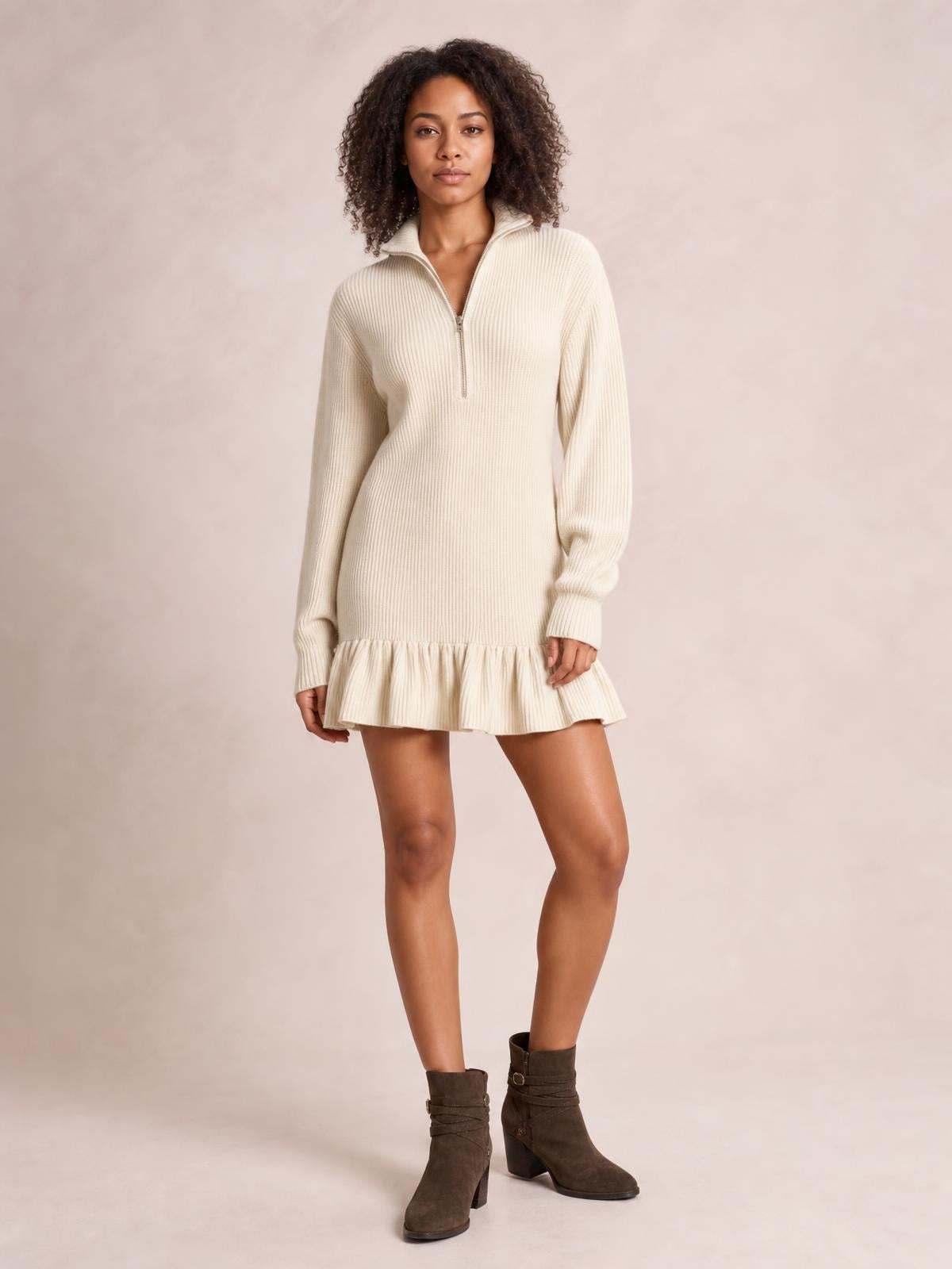 Model wears a cream ribbed half-zip dress with a high collar, long sleeves, relaxed silhouette, and playful ruffle hem while standing with one leg forward.