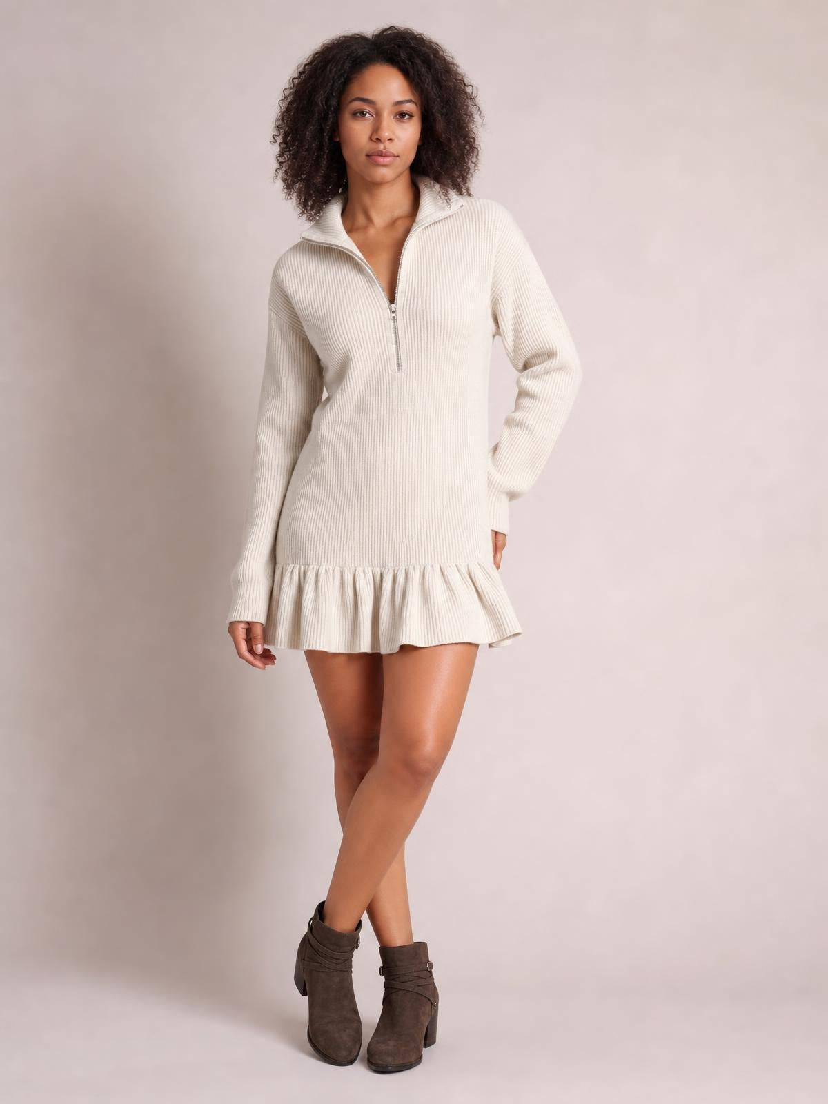 Model wears an ivory ribbed half-zip dress with a stand collar, long sleeves, relaxed silhouette, and gathered ruffle hem while posing with crossed legs.
