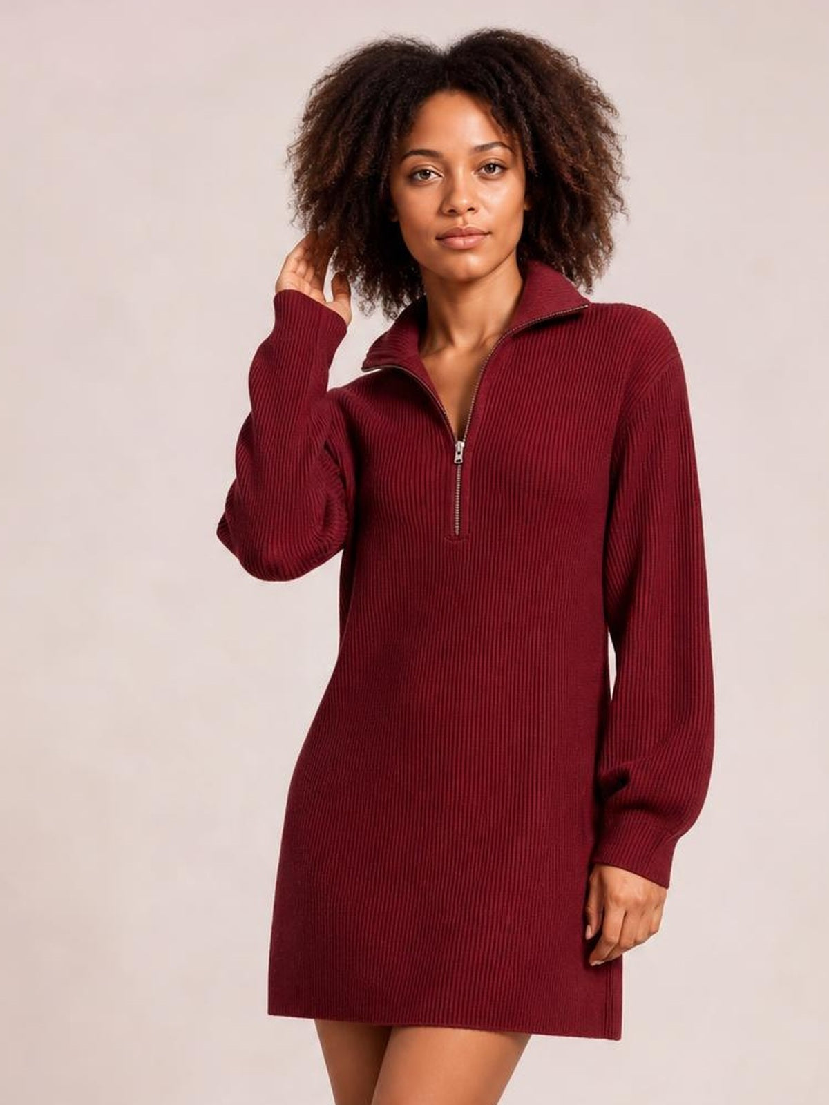 A model poses in a burgundy ribbed dress with a relaxed straight silhouette, long sleeves, stand collar, and half-zip front, lifting one hand to her hair.