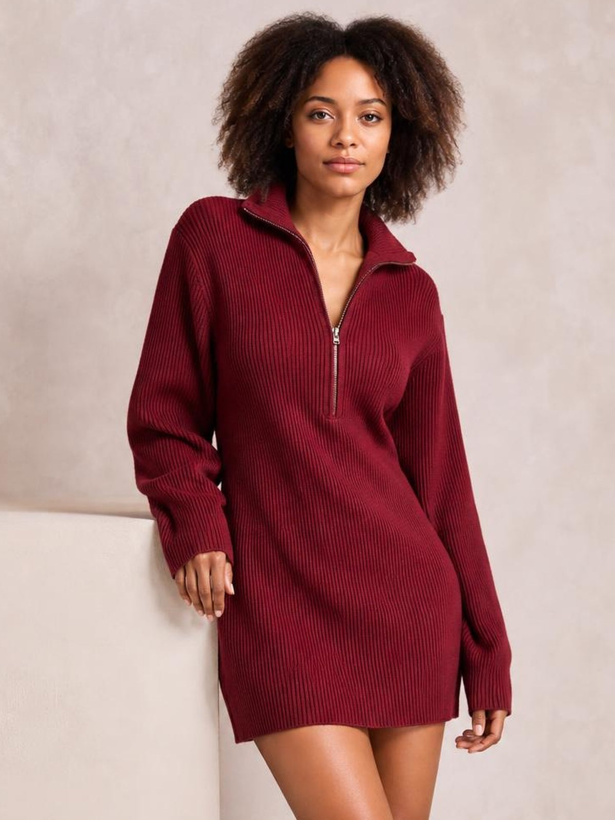 A model leans against a ledge in a burgundy ribbed dress with long sleeves, a relaxed mini-length silhouette, and a high collar with a half-zip front.