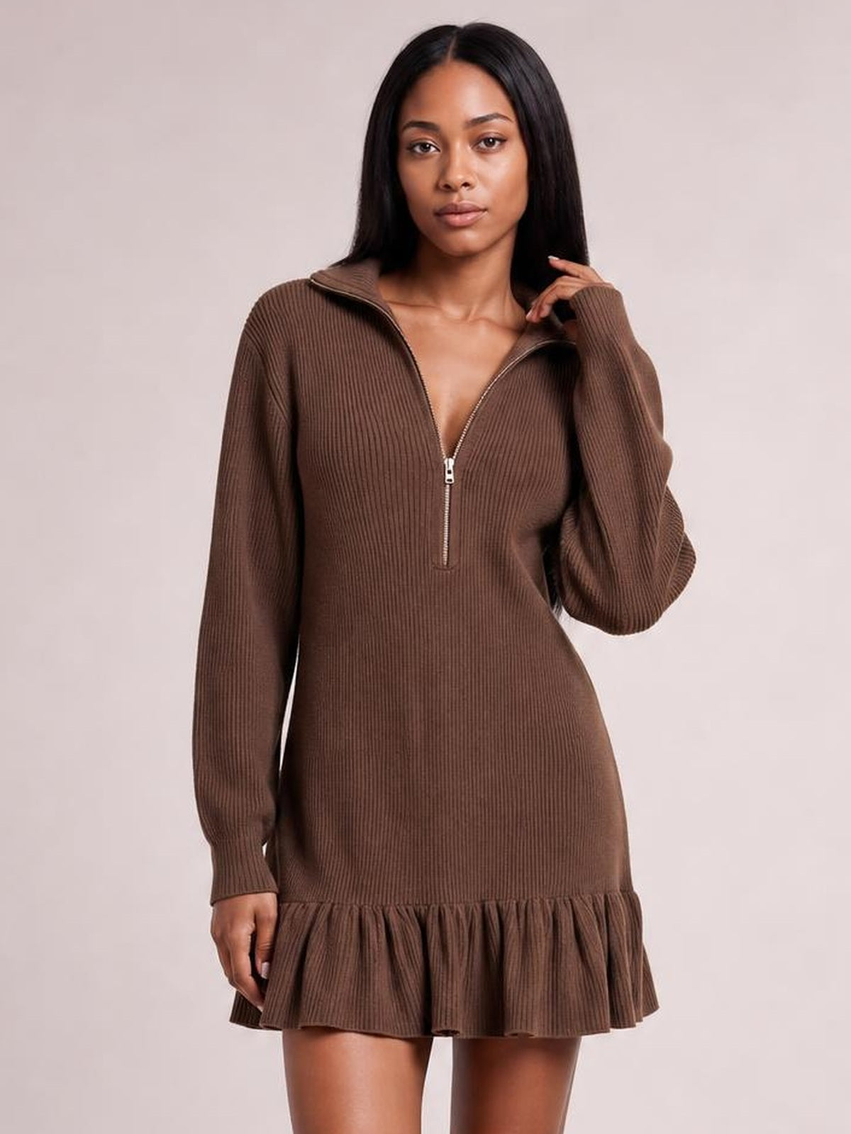 A model wears a brown ribbed half-zip mini dress with long sleeves, a relaxed silhouette, and a gathered ruffle hem, touching the collar.