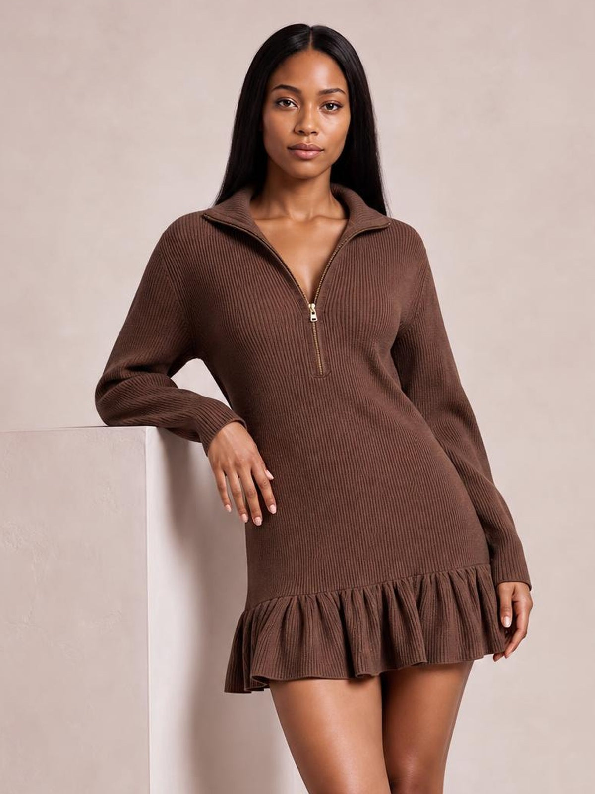 Model leans against a ledge wearing a brown ribbed half-zip dress with long sleeves, a relaxed silhouette, and a gathered ruffle hem.