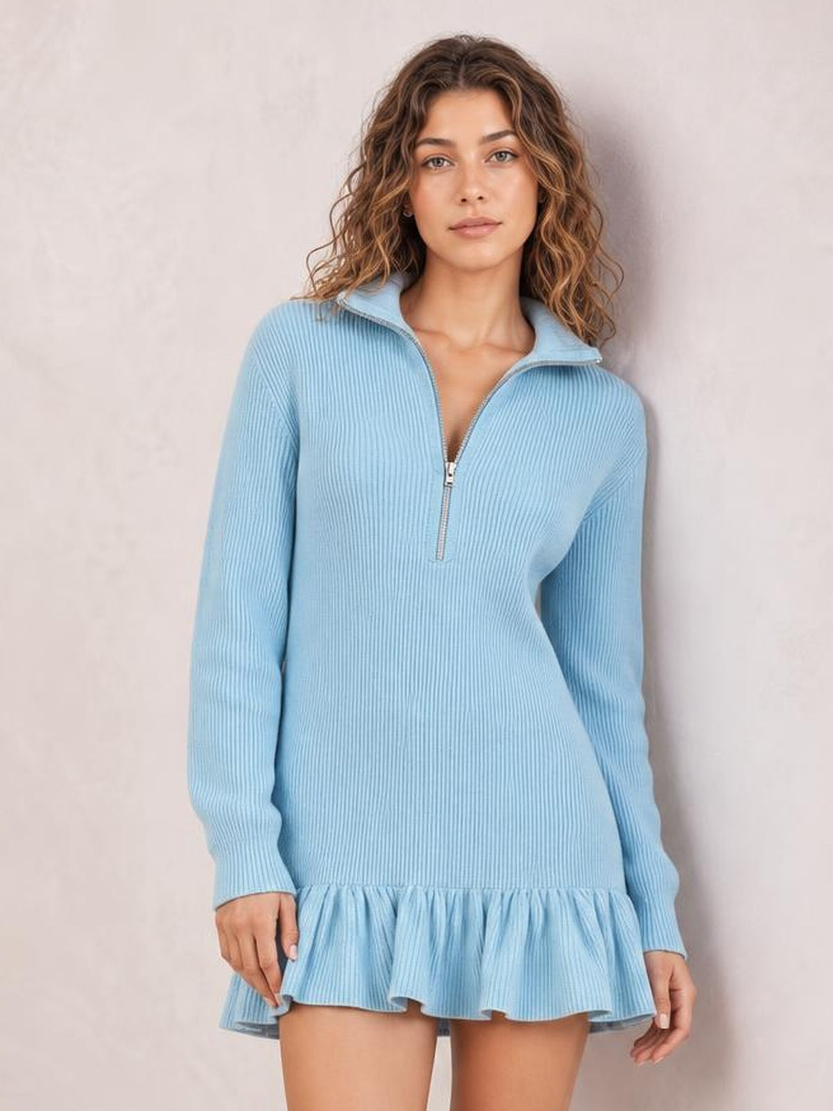 A model stands in a light blue ribbed dress with long sleeves, a half-zip collar, fitted silhouette, and playful ruffled hem.