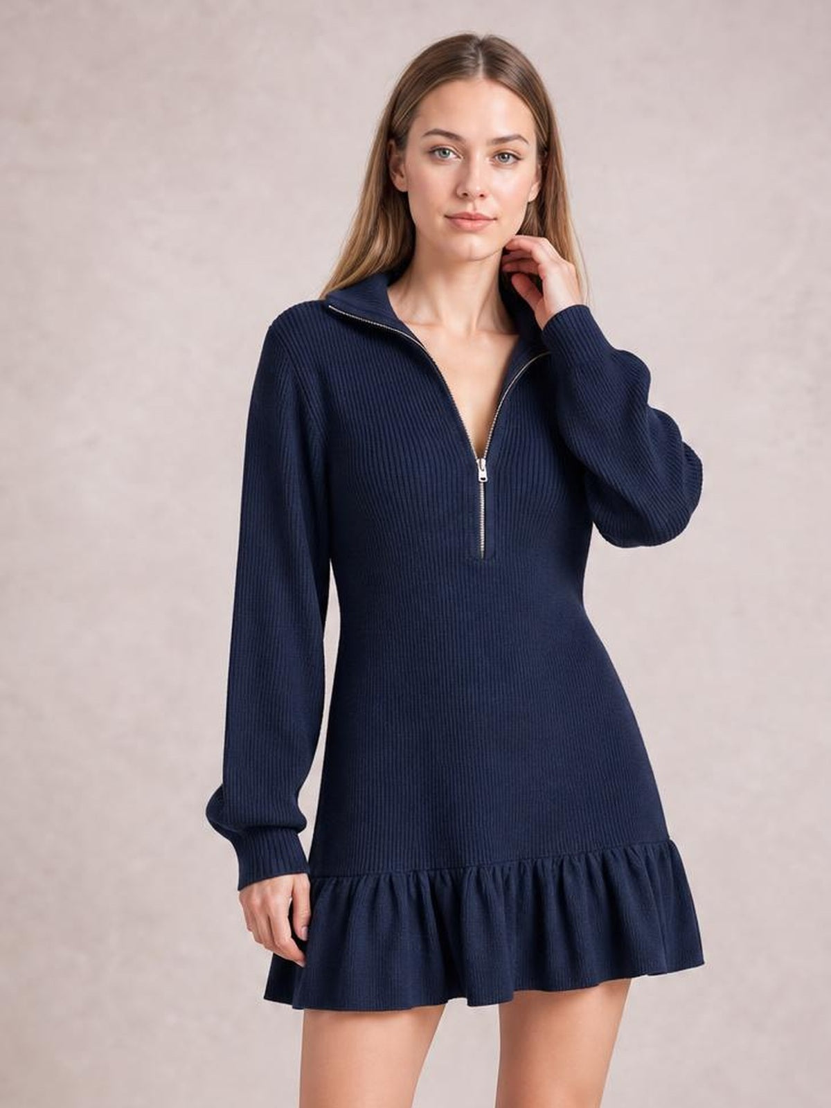 A model poses in a navy ribbed dress with long sleeves, a half-zip neckline, fitted silhouette, and flouncy ruffle hem, touching her neck.