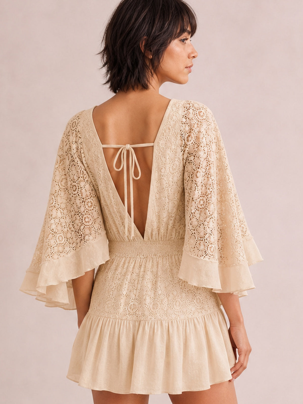 Model shown from the back wearing an ivory lace mini dress with an open tie-back, smocked waist, bell sleeves, and ruffled hem.
