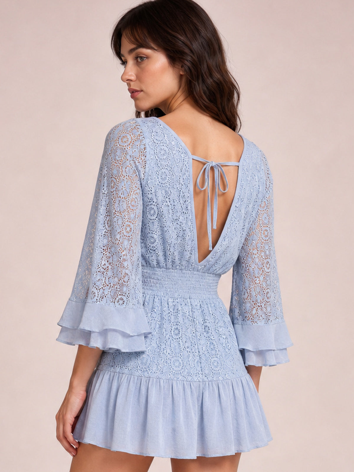 A model poses in a light blue lace mini dress with an open tie-back, smocked waist, flared silhouette, layered bell sleeves, and ruffled hem.
