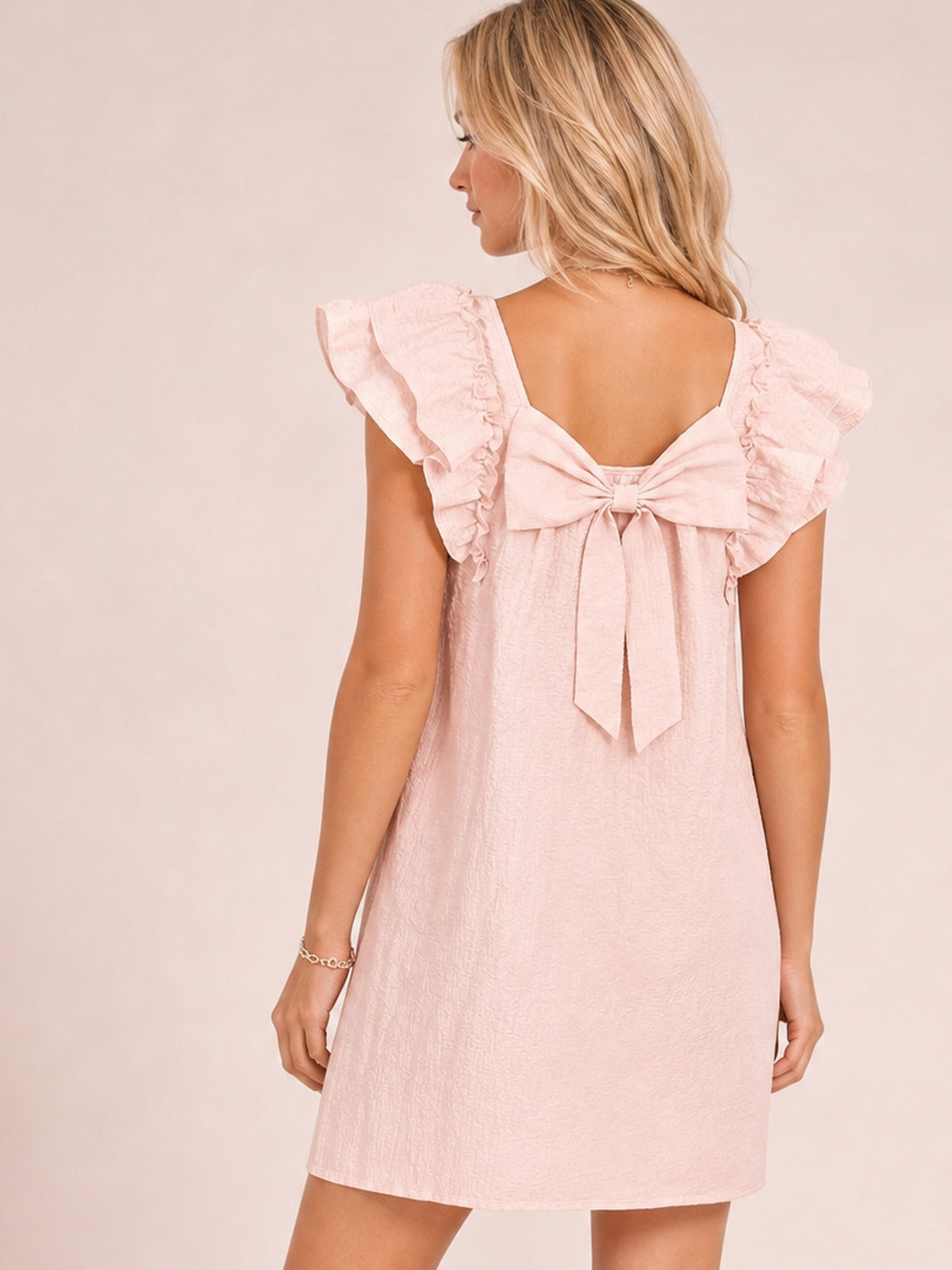 Model shown from the back in a light pink relaxed mini dress with fluttery ruffle sleeves, layered frills, and a large bow with long trailing ties.