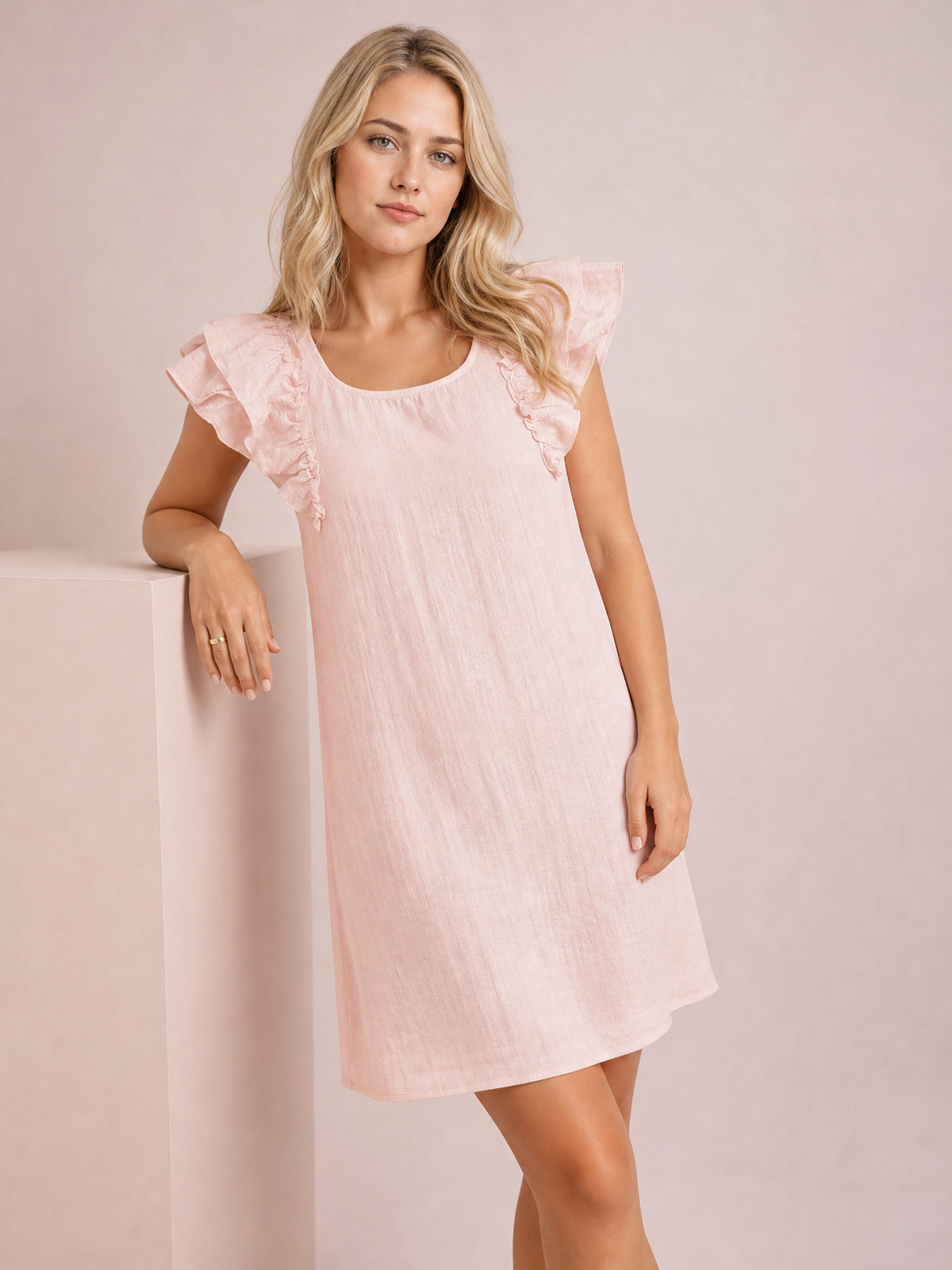 Model leans against a pedestal wearing a watermelon pink mini dress with a relaxed silhouette, rounded neckline, and fluttery ruffle sleeves.