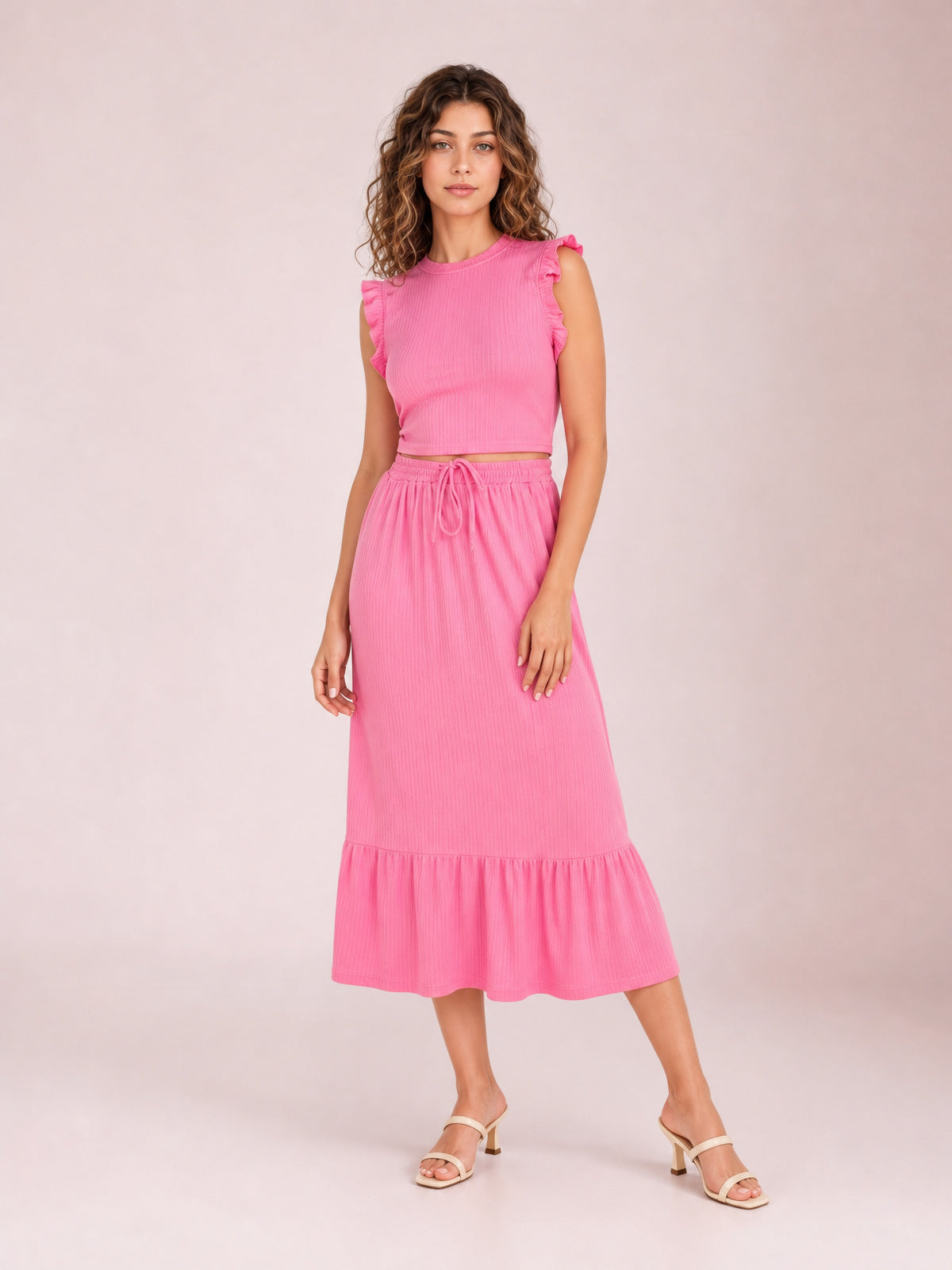 A model poses in a vibrant pink two-piece set with a round-neck ruffle-trim top and drawstring midi skirt, featuring a relaxed silhouette and tiered hem.