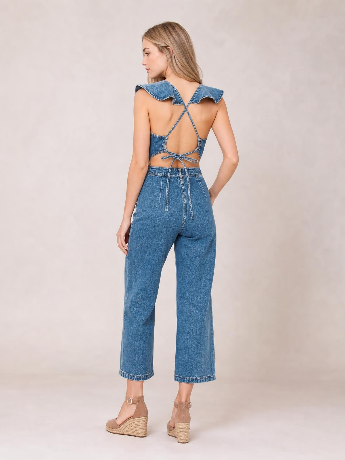 A model poses from the back in a blue denim sleeveless jumpsuit with ruffled shoulder flounces, crossed straps, an open back, tied waist, and cropped wide legs.