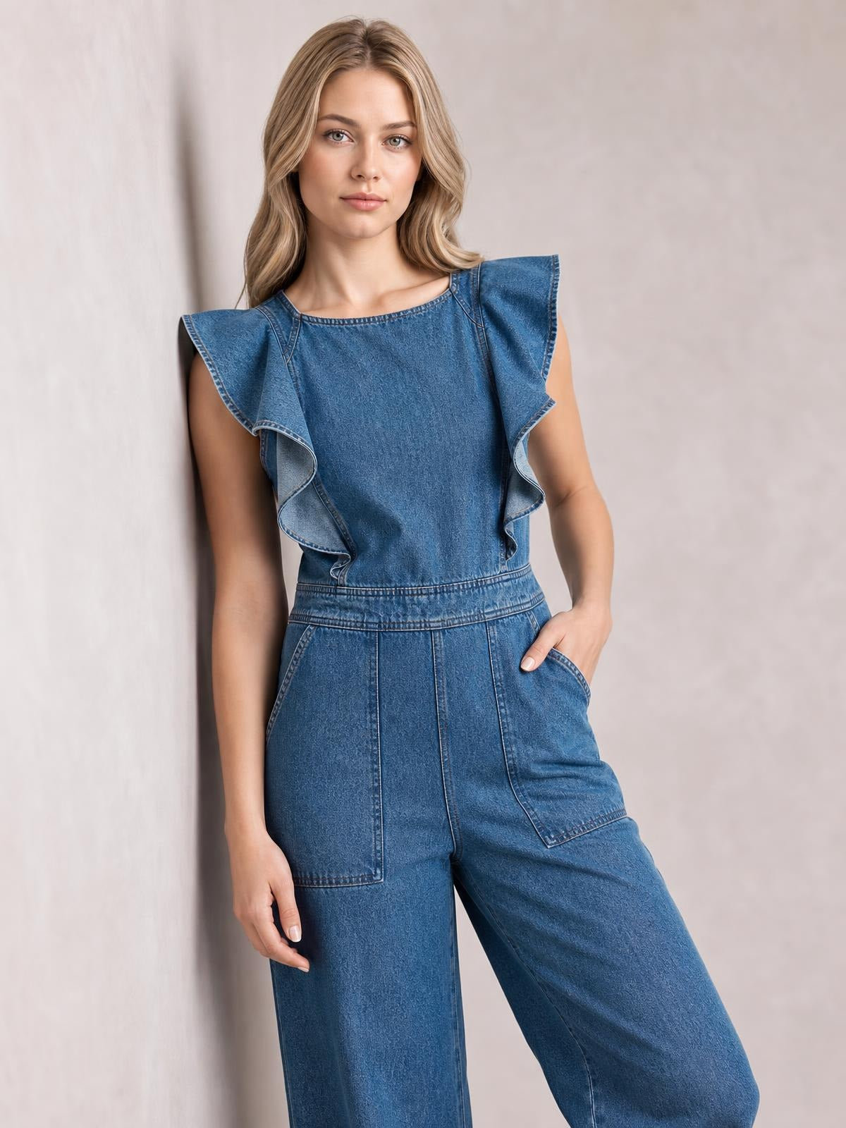 Model poses in a blue denim jumpsuit with ruffled flutter sleeves, a round neckline, defined waist, wide-leg silhouette, and large patch pockets.