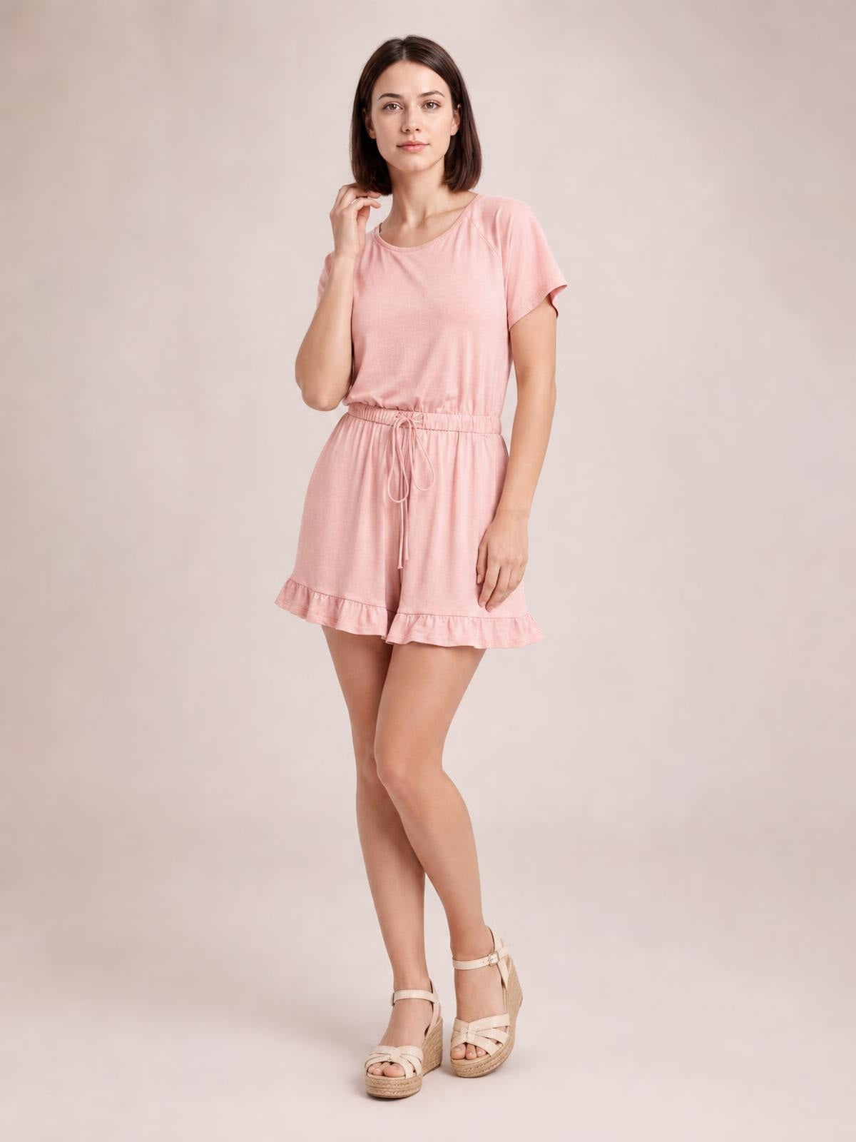 A model wears a pink short-sleeve romper with a wide boat neckline, elastic drawstring waist, relaxed shorts, and ruffled hems in a crossed-leg pose.